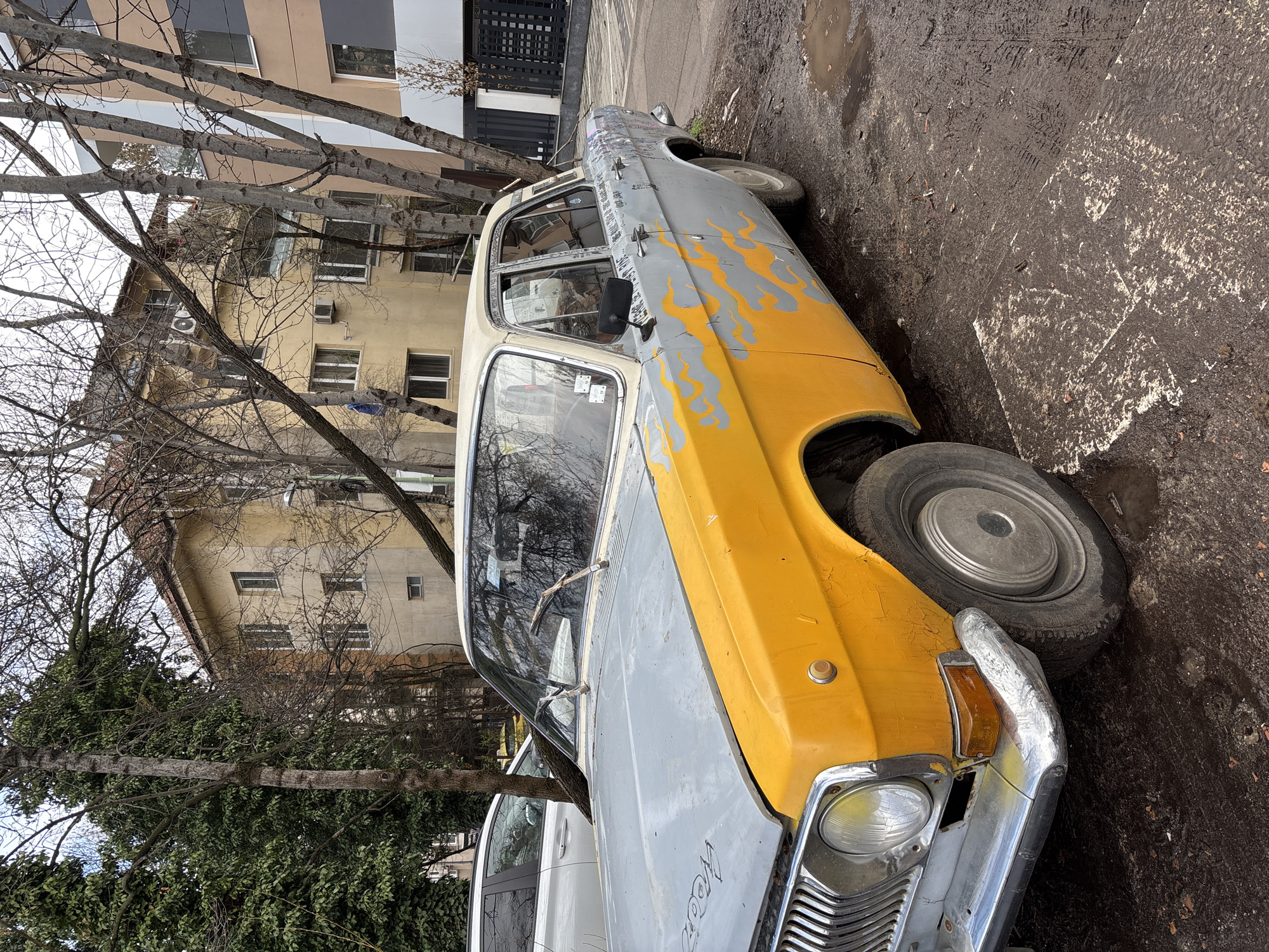 An abandoned or severely neglected yellow and gray sedan is parked on a street in Sofia, Bulgaria. The car exhibits damage and appears to be missing a wheel; another tire is present next to the front of the vehicle, suggesting a wheel change or replacement. Parts of the car are painted with flame graphics. The surrounding environment consists of a building in the background and evidence of mud or wet conditions on the pavement. Another car is parked behind the neglected sedan. Bare trees are visible in the foreground, obscuring some of the scene.