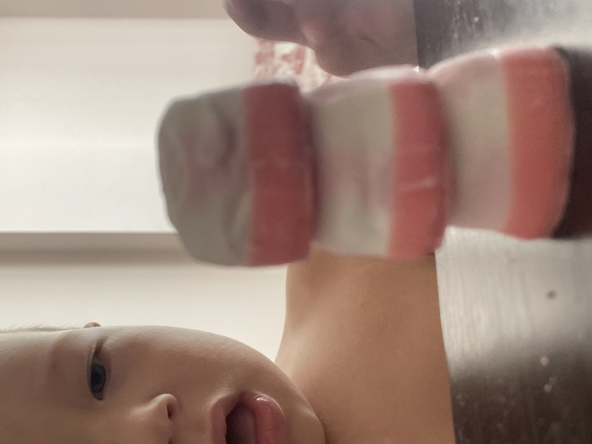 A child's face, partially obscured by a stack of three colorful candies, is visible on the left side of the frame. The candies are layered with white and pink sections. The child's mouth is open, and their eye is looking towards the candies. The background is out of focus, appearing as a light-colored wall. A hand is visible on the right side of the image, near the candies. The scene is indoors, with a wooden surface in the foreground. The location is not specified in the image or its context to be Sochi, Russia.