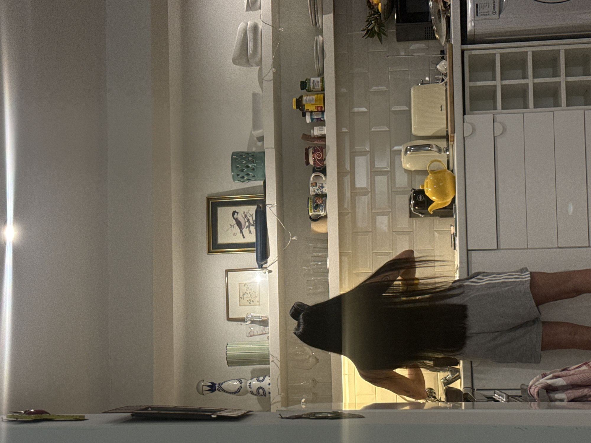 A woman with long, dark hair is standing in a kitchen. She is wearing gray shorts with white stripes. The kitchen has white cabinets, a microwave, and a kettle. There are various items displayed on shelves, including jars, mugs, and decorative objects. Artwork adorns the wall, and kitchen utensils are visible. The location is likely London, United Kingdom.