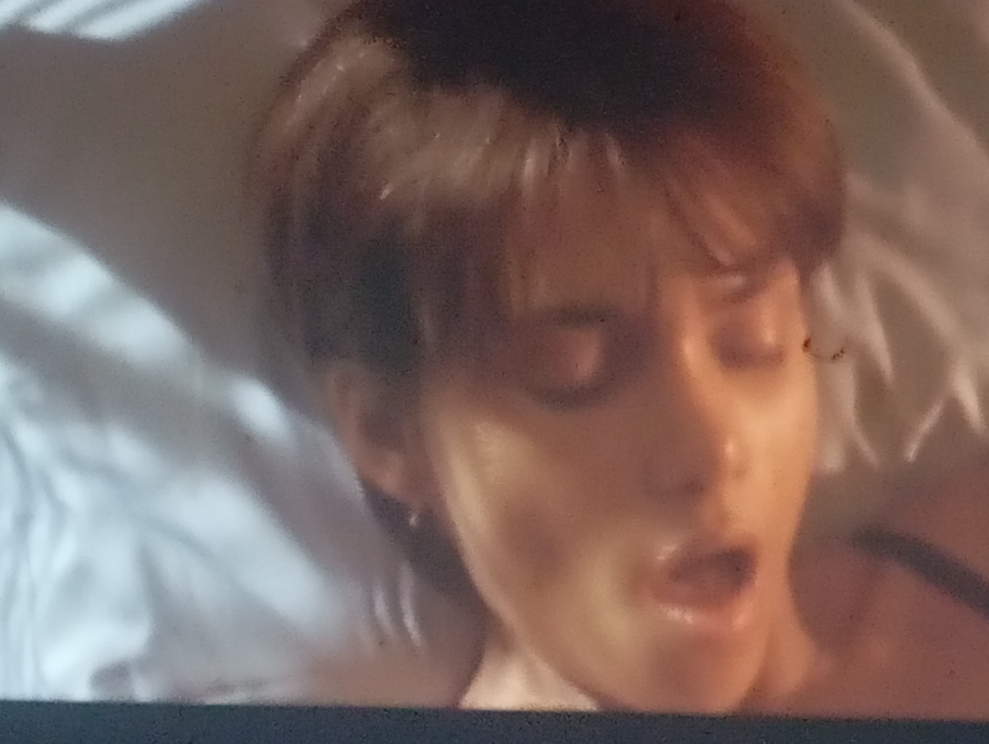 The image shows a close-up, blurry, and slightly pixelated view of a person's face and upper body, appearing to be lying down on a white, possibly bedsheeted surface. The person has short, dark brown hair and their eyes are partially closed. Their mouth is open, and their expression is somewhat ambiguous due to the low image quality. The lighting is soft and appears to be coming from the front, casting gentle shadows. There are no visible elements in the image that would suggest a specific location in Sofiivska Borshchahivka, Ukraine.