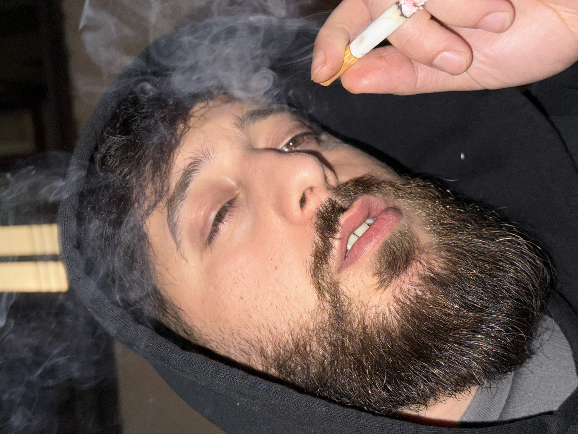 A male individual, likely in his mid-20s to early 30s, is depicted in a close-up, upward-angled shot. He has dark, wavy hair, a full, dark brown beard and mustache, and brown eyes that are partially closed or downcast. He wears a black hooded sweatshirt with the hood up, revealing a grey t-shirt collar beneath. His lips are slightly parted, revealing upper teeth. His right hand, positioned in the lower right quadrant of the frame, holds a lit cigarette between the thumb and index finger. The cigarette has a visible ash cone and glows red at its tip. Plumes of white smoke emanate from the cigarette and partially obscure the area around the individual's head, particularly to the left of his face. The background is dark and largely out of focus, with faint indications of vertical, light-colored structures in the upper left, suggesting an indoor setting. No discernible features in the image permit identification of a specific location within Strasbourg, France, or any other city.