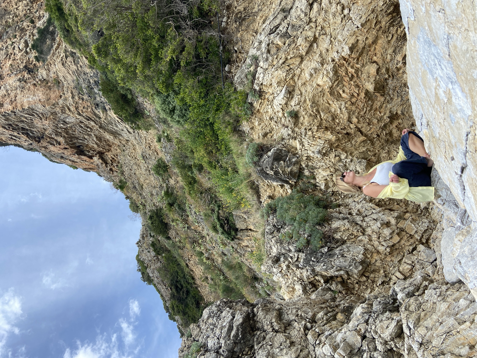 A woman with blond hair lies on a rocky cliff face in Prijevor, Montenegro. She wears dark pants, a white tank top, and a yellow garment covering her torso. The rocky terrain is composed of varying shades of tan and gray rock, with scattered patches of green vegetation clinging to the surface. To the left, the sky is visible, displaying a mix of blue and light gray clouds. The cliff face rises dramatically, extending vertically and slightly curving toward the upper left corner of the image, showcasing the geological features and vegetation. The woman is oriented horizontally, positioned on the upper-right side of the frame, seemingly sunbathing or resting.