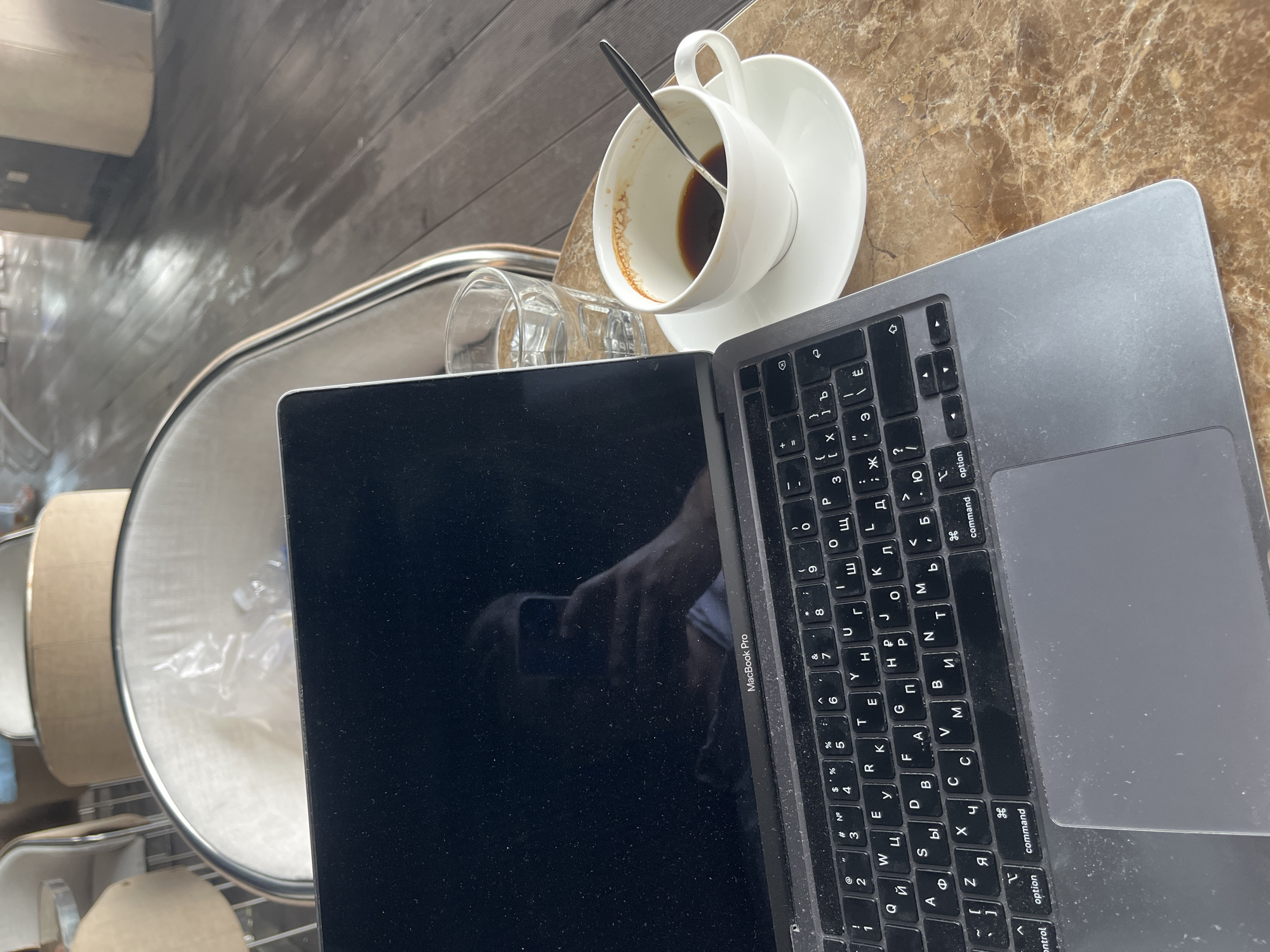The image, taken in Bac Nha Trang Ward, Vietnam, captures an overhead view of a work or study setup on a marbled brown table. A silver MacBook Pro laptop is prominently displayed, with its screen showing reflections, including a person's arm or hand holding a phone. Both the laptop screen and keyboard exhibit noticeable dust.
To the upper right of the laptop, a white ceramic coffee cup with a matching saucer sits, containing a small amount of dark liquid, likely residual coffee, and a spoon. Adjacent to the cup, a clear glass tumbler is present. The background reveals a wooden floor, and reflections on the table and laptop screen hint at the surrounding environment, possibly a cafe or an office. The overall lighting suggests it is daytime.
The laptop's keyboard features both Latin and Cyrillic characters, indicating a language layout such as Russian or Ukrainian. Visible text on the laptop includes "MacBook Pro" and various key labels like "control," "option," "command," "shift," "caps lock," "tab," "return," and "delete." Numerous Latin letters (e.g., Q, W, E, R, T) and their Cyrillic equivalents (e.g., Й, Ц, У, К, Е) are visible, along with numbers and symbols (e.g., #3, @2, $%, &7, (9, )0). The scene portrays an everyday moment of an individual engaged in an activity involving the laptop, likely accompanied by a finished beverage.