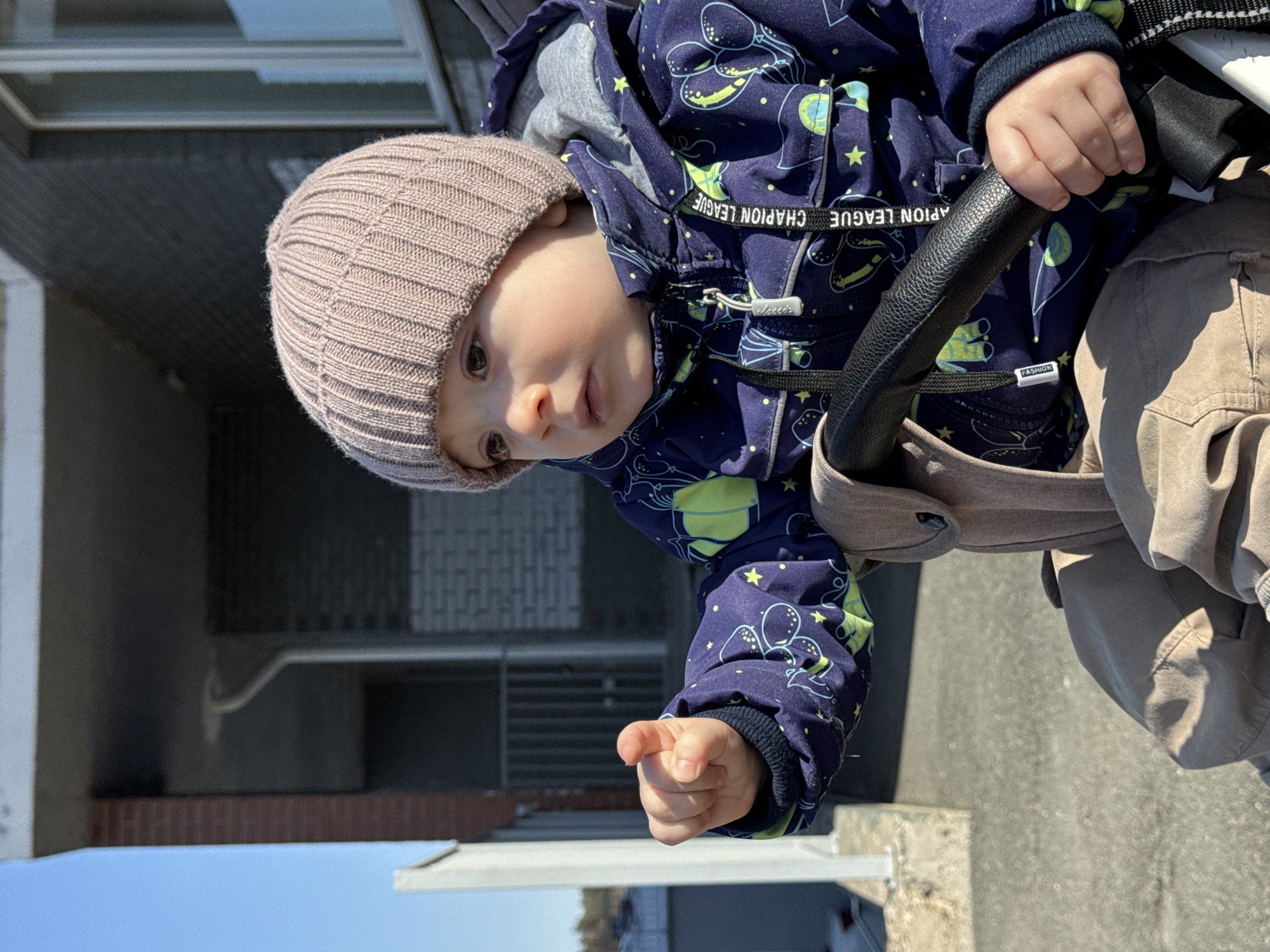 A child, wearing a light brown knitted hat and a blue jacket with yellow balloon and star patterns, is sitting in a stroller. The child is holding onto the stroller's black handlebar with one hand and pointing with the other. The background shows a building with brick and grey concrete elements, and a glimpse of blue sky. The location is Izhevsk, Russia.