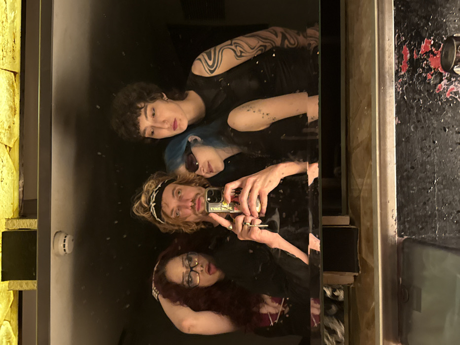 The image is a mirror reflection of four individuals in a dimly lit indoor setting, likely a social venue in Tel-Aviv, Israel; the exact location is not discernible. The background features dark walls and ceiling, with a prominent textured golden ceiling fixture and a white smoke detector visible overhead. Below the mirror, a dark, textured counter or surface displays reddish residue and a metallic object on the right.
From left to right, the reflected individuals are:
1. A woman with long, curly reddish-brown hair, dark-rimmed glasses, and a dark top. Her left hand is raised, with fingers near her head, and her mouth is open in an expressive pose.
2. A man with light brown, shaggy hair, a dark headband featuring a white barbed wire pattern, a light mustache, a soul patch, and a nose ring. He wears a dark, textured long-sleeved shirt. He holds a white smartphone, which has a colorful sticker on its back, in both hands, angled to capture the reflection. A set of keys hangs from his left hand.
3. A woman with light blue hair, dark, narrow sunglasses, and a dark sleeveless top. Several small dark marks are visible on her left arm. She displays a subtle smile, with her head slightly tilted.
4. A woman with short, dark, curly hair and a dark sleeveless top. Her left arm is extensively tattooed with abstract, wavy black patterns. She maintains a neutral expression.
The group is captured posing together, appearing to take a mirror selfie.