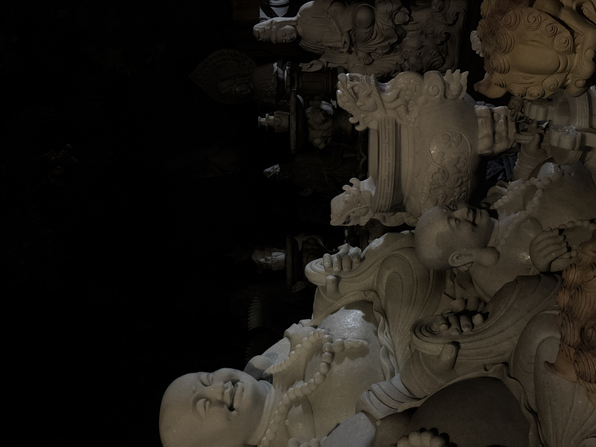 The image shows a collection of stone sculptures, likely depicting Buddhist figures and mythical creatures such as dragons and lions. The sculptures are densely packed together, and the lighting is dim, emphasizing the intricate details of the carvings.
These sculptures are likely located in a temple or cultural site in the city of Đà Nẵng, Vietnam. Specifically, they may be found in the Marble Mountains area, known for its caves, temples, and stone carving workshops.