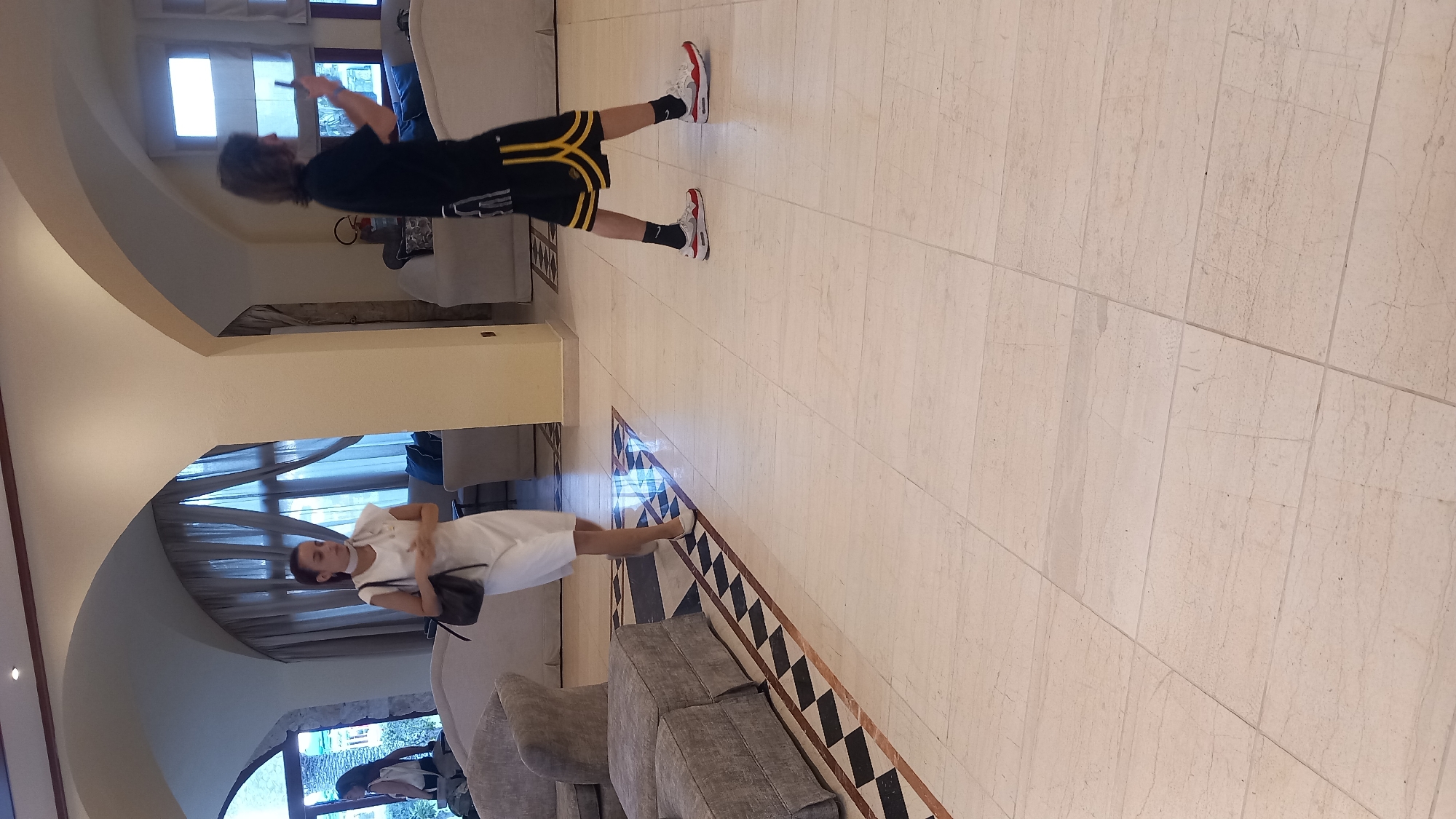A full shot of a hotel lobby in Kos, Greece, shows two individuals walking on a marble tiled floor. A woman on the left, dressed in a white dress and carrying a black purse, walks towards the viewer. To her right, a person wearing a black t-shirt, black shorts with yellow trim, and white sneakers walks away from the viewer, holding a phone. The lobby features cream-colored arched ceilings, large windows with draped curtains, and plush seating areas with sofas and armchairs. Decorative black and beige tiling is visible along the floor.