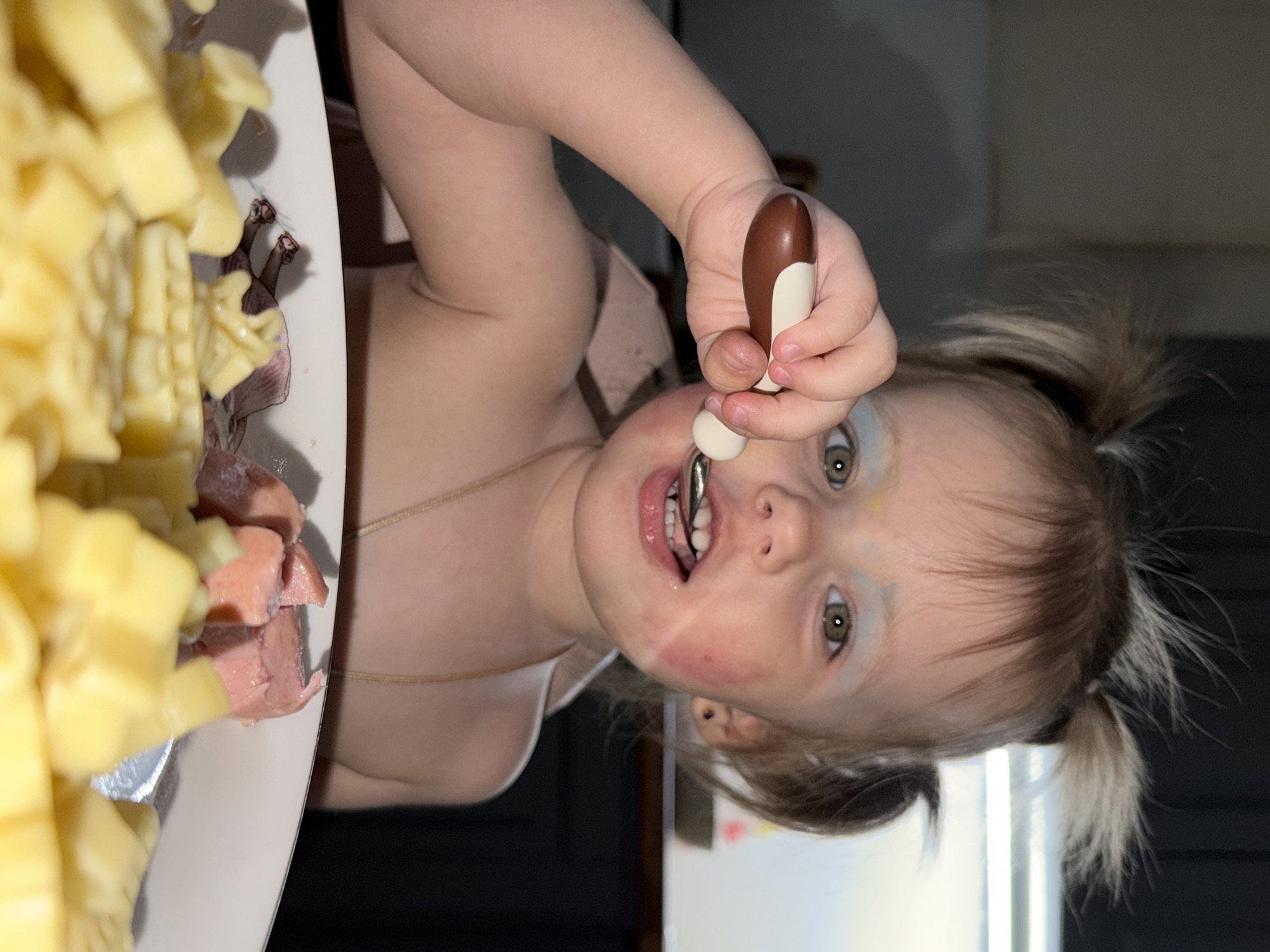 A fair-skinned toddler with hazel eyes and blonde hair styled in pigtails is eating from a white plate containing diced cheese and sausages. The child holds a spoon in their right hand, placing it in their mouth. Traces of red coloring, possibly makeup, are visible on the child's cheek. The background is a dark room with a white wall or door. The scene is presumably indoors, but the location in Reshetnikovo, Russia is not apparent from the image.