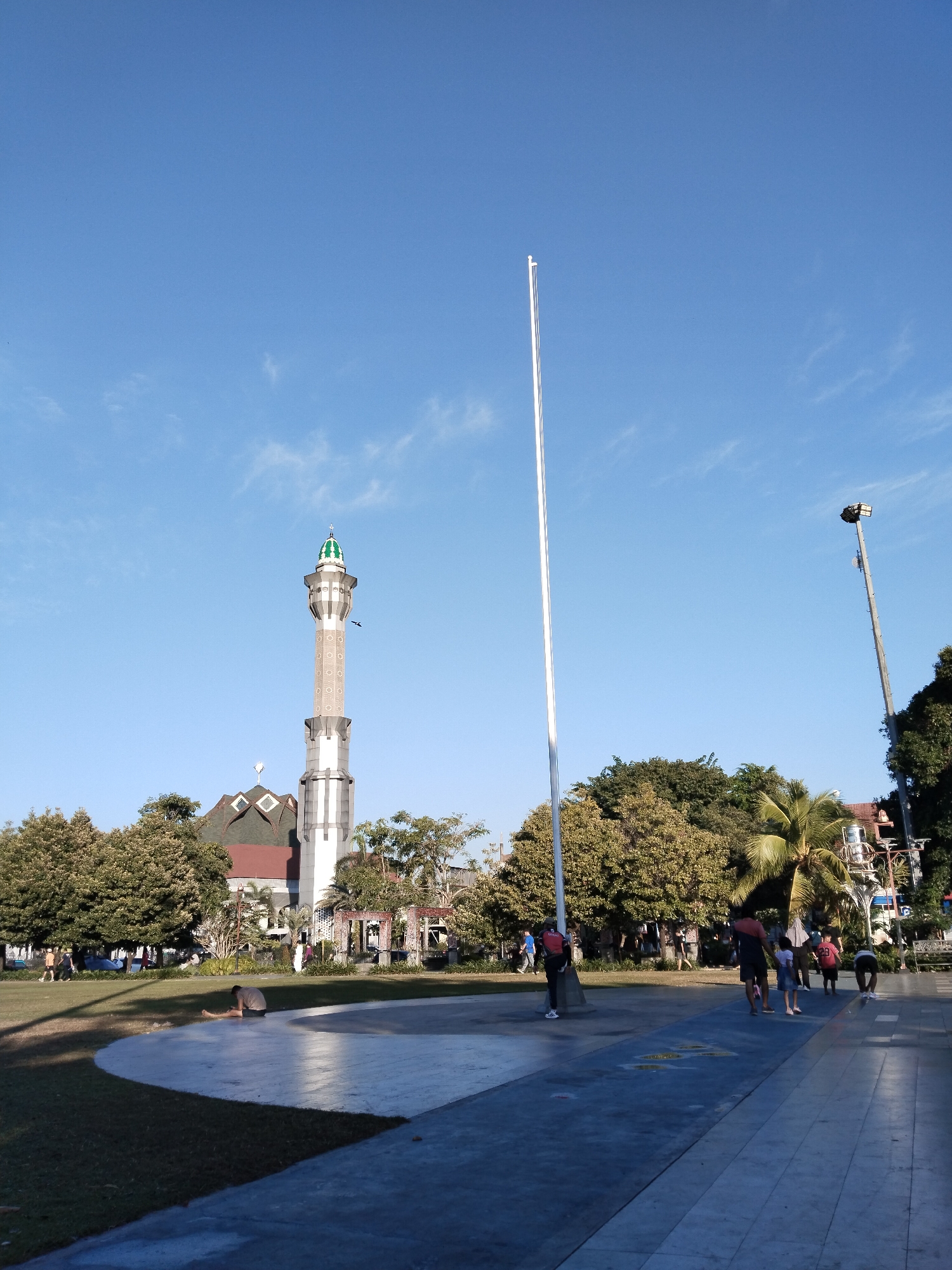 This image captures a daytime scene in a public park area in Kecamatan Pati, Indonesia. A tall, slender flagpole stands prominently in the foreground, centered within the frame and reaching towards a clear blue sky. To the left, a distinctive mosque with a tall, ornate minaret and a green-capped roof is visible, surrounded by lush green trees. In the middle ground, several people are scattered across the park. Some are walking along a paved pathway, while others are engaged in activities on the grass. One individual is seated on the ground near the pathway, appearing to stretch or rest. The park features a mix of grassy areas and paved surfaces, with additional trees and some architectural elements like a trellis structure in the background. The lighting suggests it is late afternoon, with long shadows cast across the scene.
