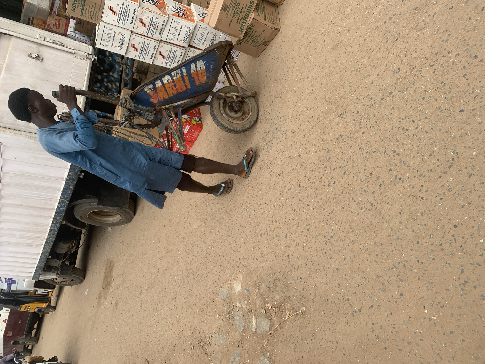 The image shows a young man in a blue traditional tunic and shorts, wearing flip-flops. He is standing next to a handcart with one wheel, holding onto its handles as if to push it. The handcart is blue and orange with the word "Targo" visible on its side. Behind the man and handcart, there are stacks of boxes, some labeled "SUNSHINE air freshener" and "SIPERCO NIGERIA LIMITED 250ml x 24pcs." The scene appears to be outdoors on a paved surface, likely a street or market, with a large white truck visible on the left side of the frame. The lighting suggests it is daytime. The man's posture and grip on the handcart imply he is about to move the cart or is in the process of loading or unloading it. The presence of the truck and stacked boxes suggests a commercial or logistical setting.