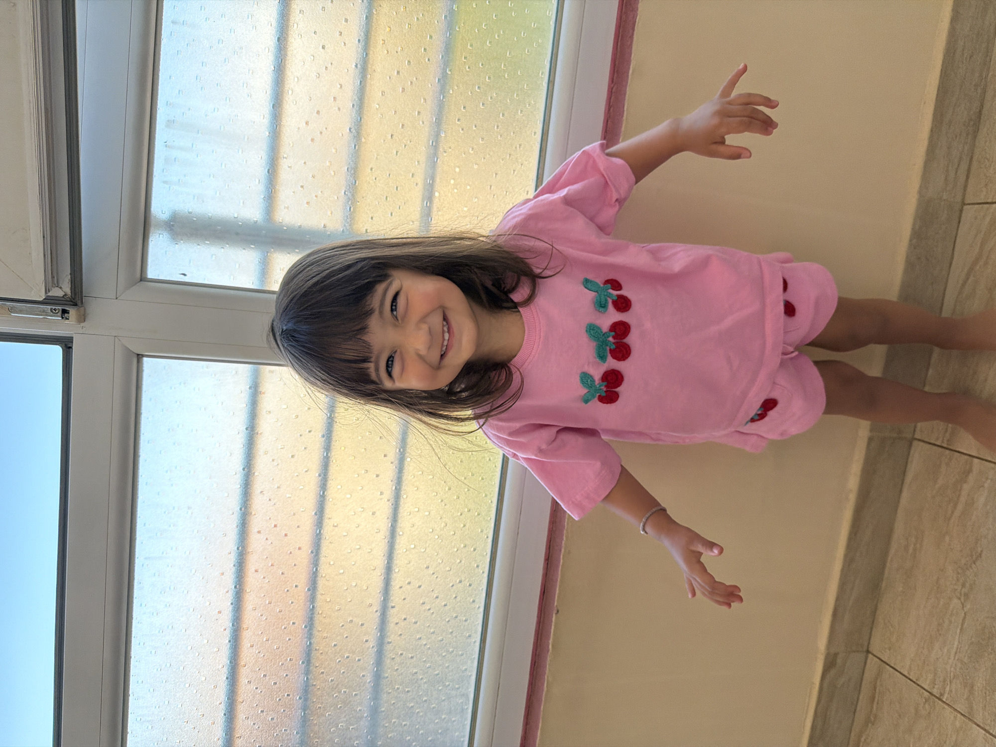 A young girl with dark hair and bangs, wearing a pink t-shirt with embroidered cherries and matching pink shorts, stands in front of a window. The window is divided into panes with a textured, semi-transparent film, allowing diffused sunlight to pass through. The girl is smiling and looking at the camera with her arms outstretched. She has a bracelet on her left wrist. The floor appears to be tiled. The background wall is a light beige color with a horizontal pink trim near the top of the wall. The scene is indoors, likely in a room with a window overlooking an outdoor area. This scene does not provide enough visual information to determine the specific location in Burgas, Bulgaria.