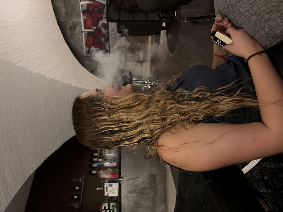 An indoor scene in San Bernardino Tlaxcalancingo, Mexico, captures a woman in profile, seated and exhaling a plume of white vapor from her mouth. The woman, with long, wavy, blonde and light brown hair, wears a dark blue sleeveless top. She holds a light-colored, rectangular vaping device with black accents in her left hand. A dark bracelet is on her right wrist, and a red string bracelet on her left. The background features a dim interior with architectural details including a white, vaulted, exposed brick ceiling and dark, textured walls. To the left, a bar area is visible with shelves holding various bottles and a box labeled "AMSTEL ULTRA." To the right, two framed artworks with abstract red and black shapes hang on a dark wall above dark seating. The woman is seated on what appears to be a dark leather sofa or chair, with a gray fabric surface beneath her left arm.