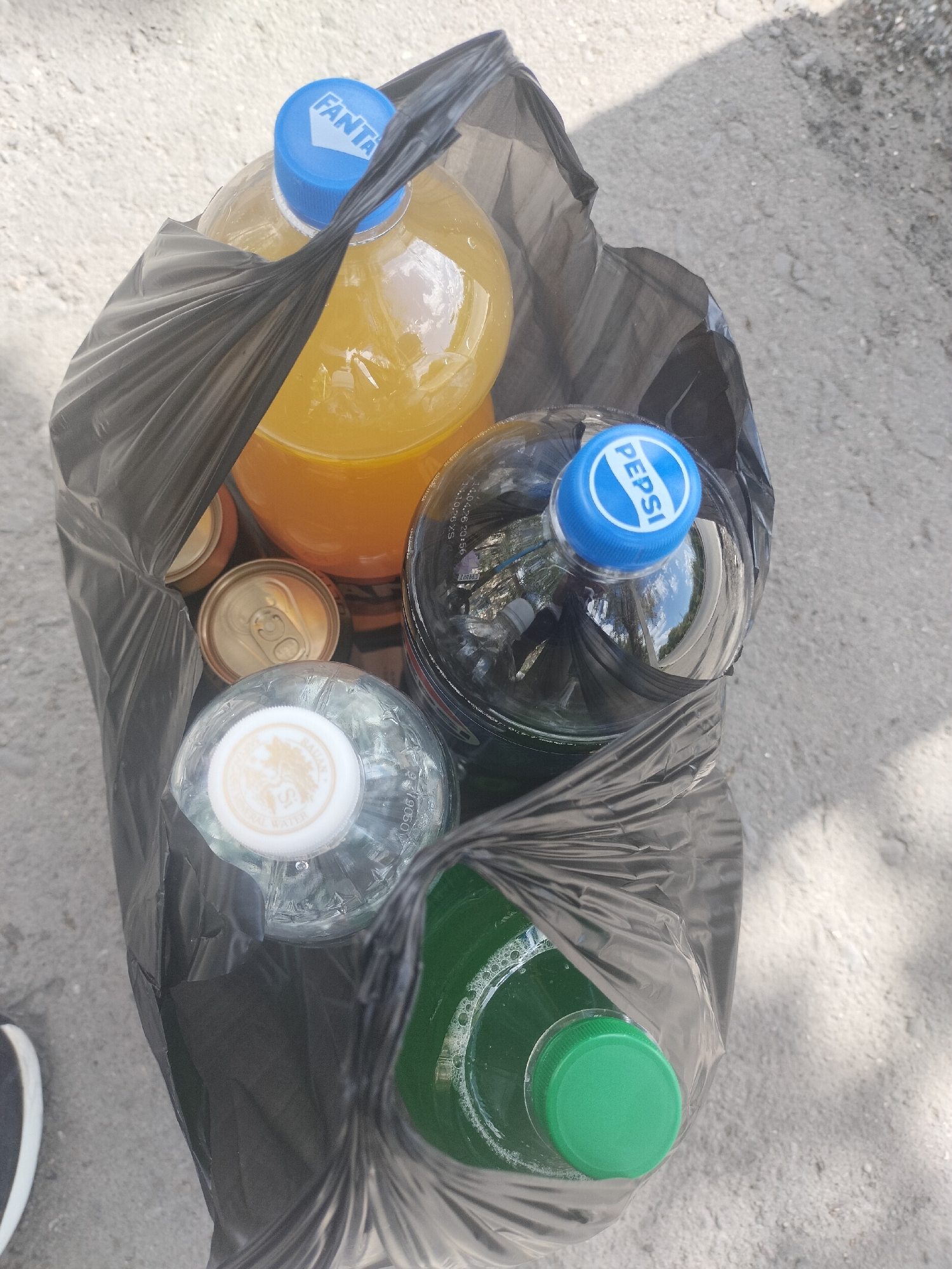 A black plastic bag is filled with various beverages and sits on a paved surface. The bag contains a large bottle of Fanta with a blue cap, a bottle of Pepsi with a blue cap, a clear bottle of water with a white and gold cap, a can of an unidentified beverage, and a green bottle with a green cap. The background is a grey, rough textured pavement with scattered light and shadows. The image is taken from a top-down perspective.
