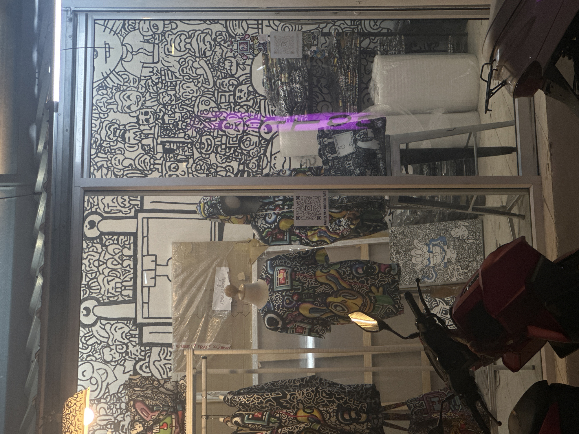 View of a shop display in Baan Lamai, Thailand. The shop window is divided into two sections, showing mannequin torsos wearing patterned shirts. The backdrop features black and white doodle art. QR codes are affixed to items. A motorcycle partially obstructs the view on the right side of the image. The light conditions suggest either evening or indoor lighting. The display includes a variety of items wrapped in plastic.
