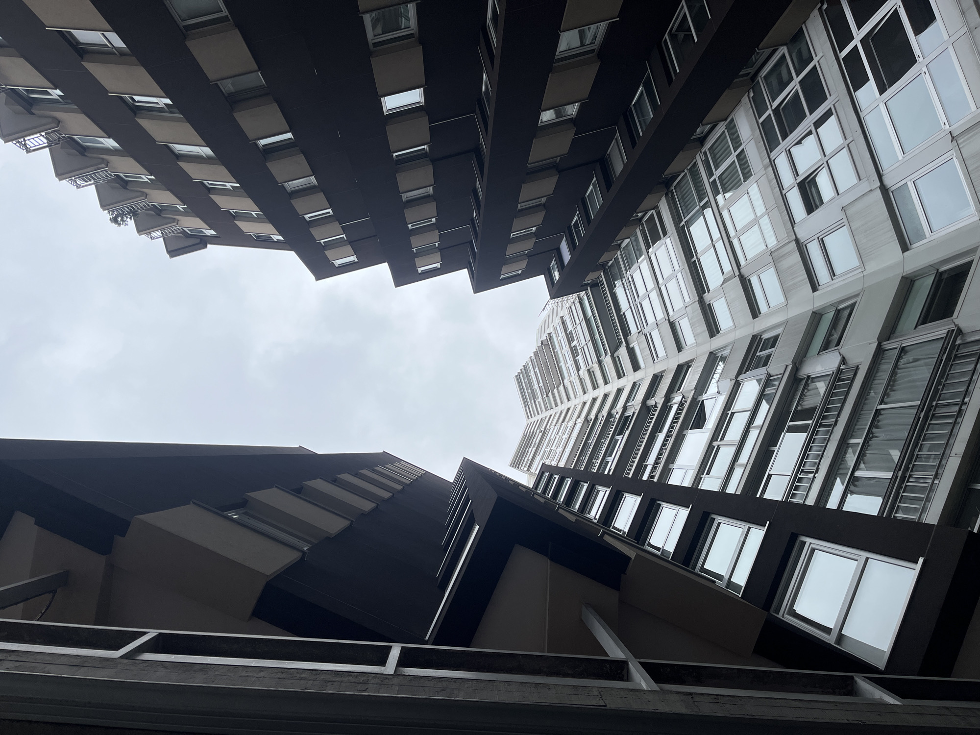 Looking upwards, the image captures the architecture of buildings in the city of La Coruña, Spain. Two buildings of varying architectural styles occupy most of the frame. The building on the left exhibits balconies and dark brown-colored construction, while the one on the right is a lighter gray with large windows, and what appears to be fire-escape ladders. The sky above is overcast, creating a muted light. The bottom edge of the frame includes a gray, metal framework.