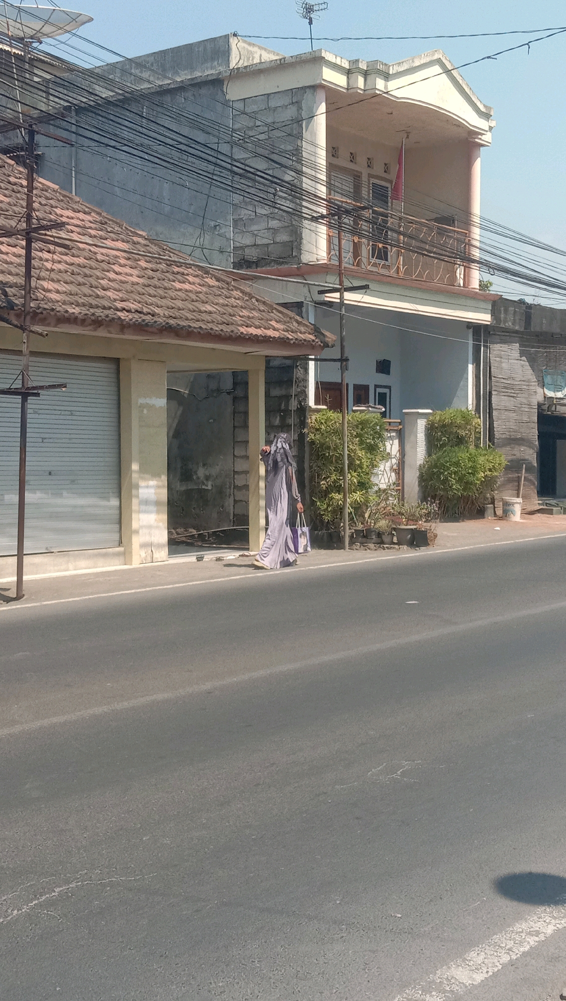 A full shot captures a pedestrian in a long, light purple garment and headscarf walking on the sidewalk next to a road in Karang Anyar, Indonesia. The pedestrian is carrying a large, light purple bag in their right hand. The sidewalk is lined with potted plants and small trees. Behind the sidewalk is a row of buildings. The building on the left has a tiled roof and a metal roll-up door. The building on the right is a two-story structure with a balcony on the upper level. Overhead, numerous electrical wires crisscross the scene, supported by poles. The road in the foreground is asphalt with faint white markings. The sky is clear and blue. The scene is bathed in sunlight, casting shadows on the road.