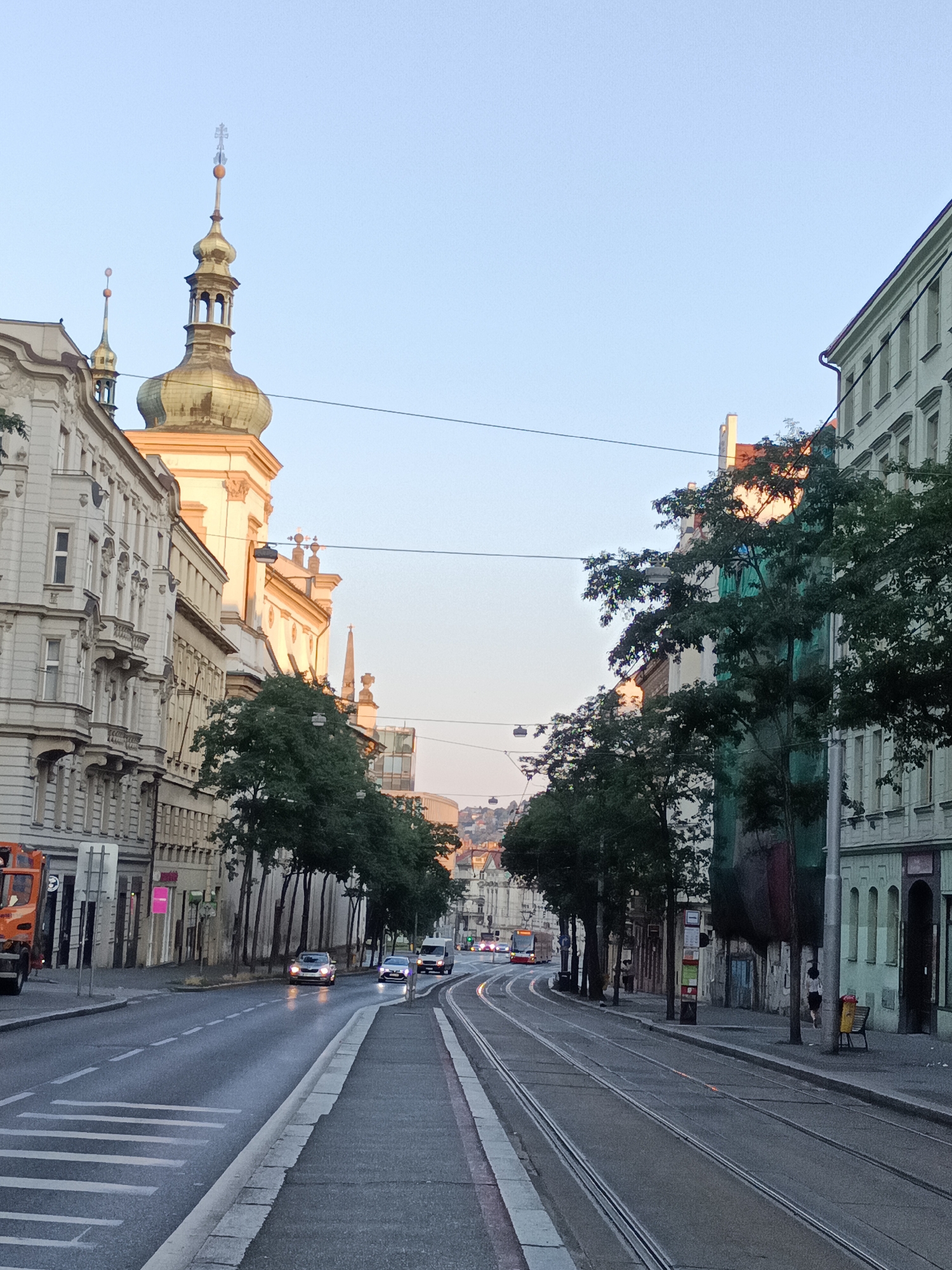 The image depicts a street scene in Prague, Czechia, likely during early morning or late evening due to the soft lighting. The perspective is from a street-level viewpoint, looking down a road with tram tracks. On the left, ornate historic buildings line the street, featuring balconies and detailed facades. Prominently visible is a building with a golden dome and spire, characteristic of Prague's architecture. Several trees with dense green foliage are planted along the sidewalk. The road itself is wet, suggesting recent rain or cleaning, with white lane markings and a crosswalk visible. Vehicles, including cars and a tram, are present on the road, some moving and others stationary. On the right side of the street, more buildings are visible, some appearing older with arched windows and others more modern. A woman in dark clothing is walking on the sidewalk. The sky above is clear and pale blue, transitioning to a warmer hue near the horizon. Power lines crisscross the upper portion of the image. In the distance, hills with buildings can be seen. The overall impression is of a quiet urban environment in a historic European city.