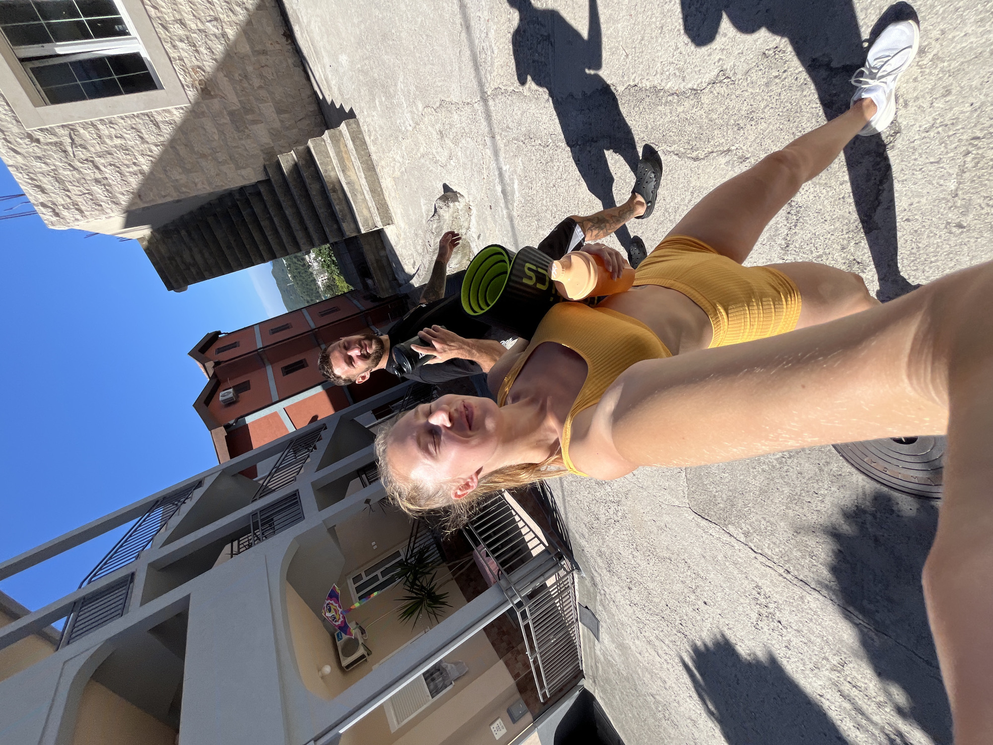 In Boreti, Montenegro, a young woman with blonde hair pulled back, wearing a yellow sports bra and matching shorts, is taking a selfie. She is holding a light orange water bottle in her right hand. Behind her, a man with short brown hair and a beard, wearing a black t-shirt and shorts, is walking. He is carrying a rolled-up black yoga mat with green accents and a black shaker bottle. They are walking on a paved surface with shadows cast by buildings and trees. In the background, there are multi-story buildings with balconies and staircases, and a clear blue sky. The scene appears to be a sunny day in a residential or resort area.