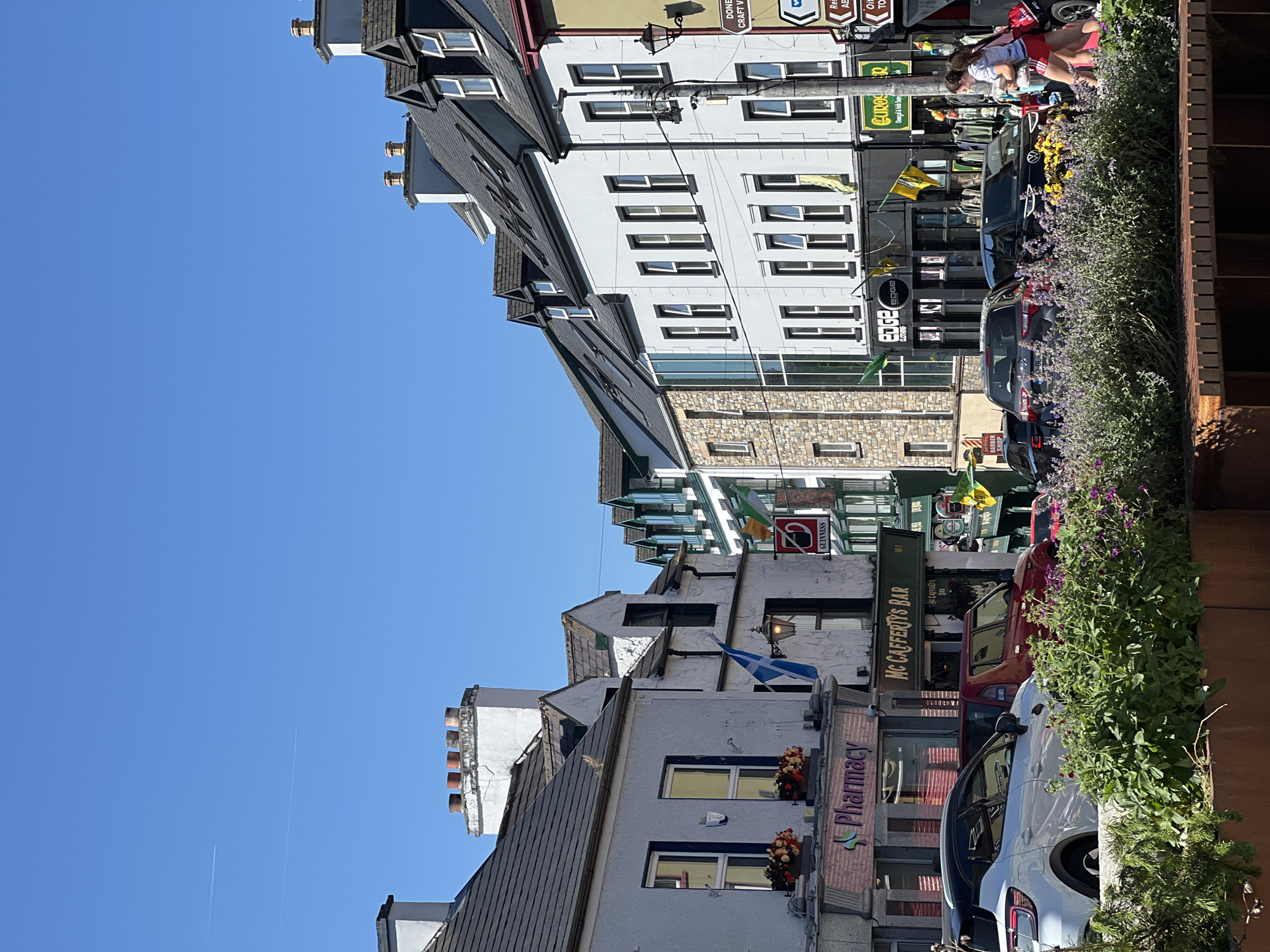A street scene in Donegal, Ireland, depicts a row of buildings with businesses on the ground floor and residences above. The sky is clear and blue with contrails from an airplane visible in the distance. The buildings are varied in style, with some featuring white stucco facades and dark tiled roofs, while others have stone accents. Several flags, including a Scottish flag and Irish flags, are displayed from the buildings.
Businesses visible include "Pharmacy," "McCafferty's Bar," "Edge Clothing," and "Eurocraft." Cars are parked along the street, and a white SUV is in the foreground. People are walking on the sidewalk, with one woman in red shorts and a t-shirt carrying bags. Flowers and greenery are planted in planters along the edge of the sidewalk. A street lamp is visible on the right side of the image, along with directional signs. The overall impression is of a sunny day in a lively town center.