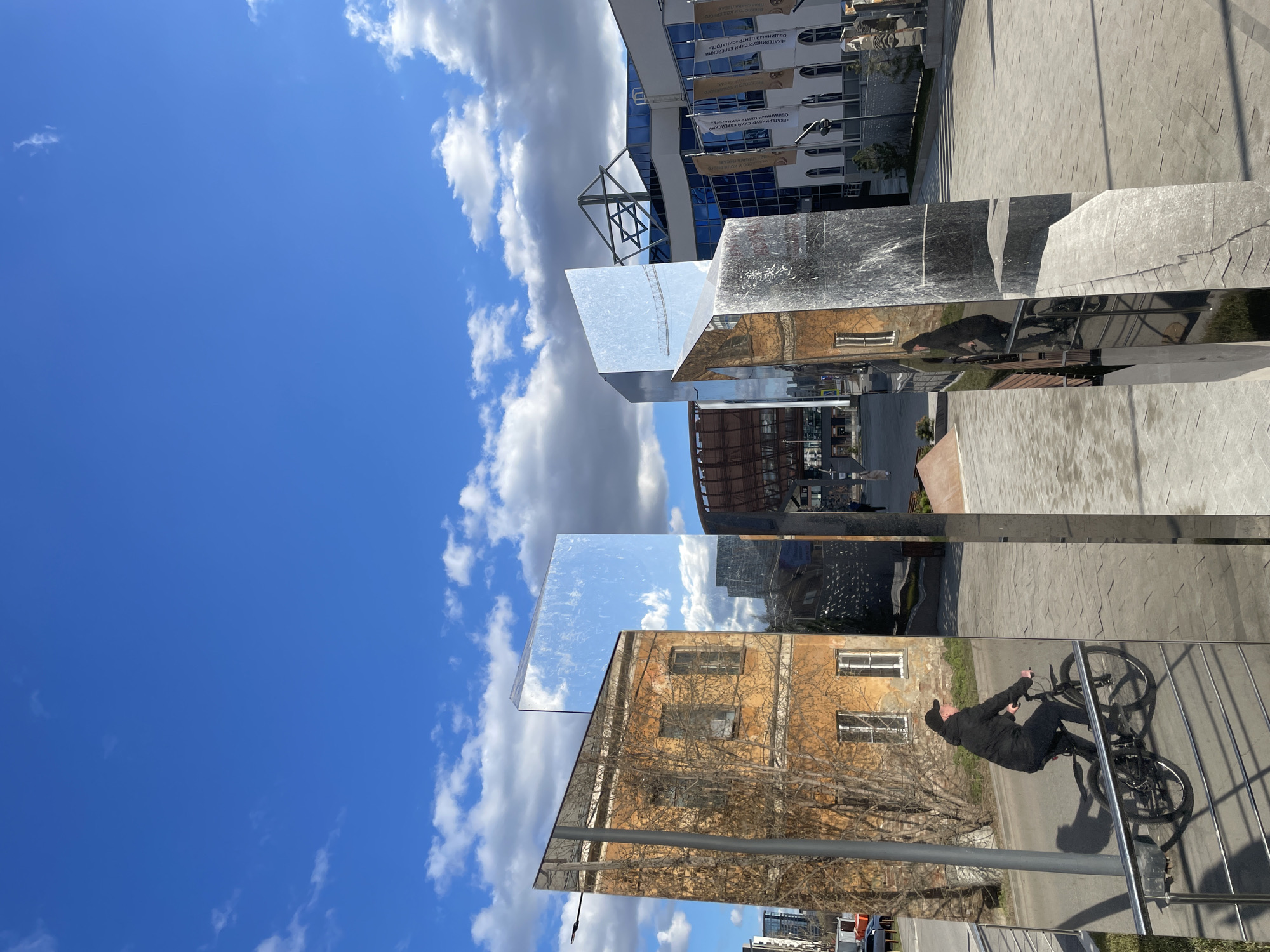 The image shows a vertical, medium shot taken in Yekaterinburg, Russia, focusing on mirrored sculptures reflecting the surroundings. The sky is bright blue with scattered white clouds. A building with a yellow facade and leafless trees is mirrored in the lower sculpture, along with the reflection of a person riding a bicycle. The person is wearing dark clothing and a cap. The upper sculpture reflects a modern building with blue glass and signage in Russian. A Star of David can be seen on the building. The ground appears to be paved with grey concrete blocks.