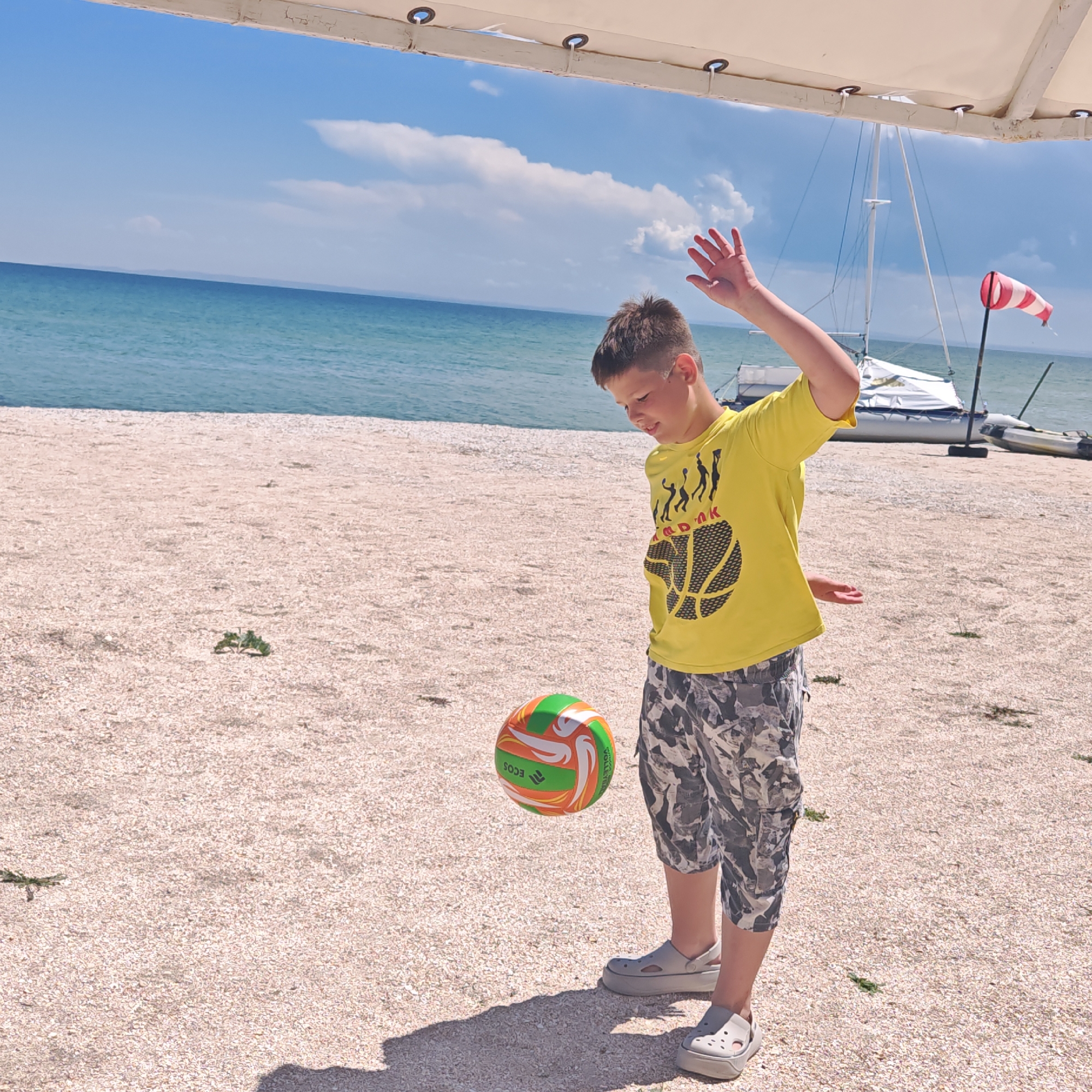 A young boy in a yellow t-shirt and camouflage shorts is playing with a volleyball on a pebble beach. He is looking down at the ball, which is in mid-air, and appears to be about to hit it with his hand. The boy's right arm is raised, and his left hand is extended downwards. In the background, the sea is visible, with a sailboat and a windsock on the shore. The sky is blue with some clouds.