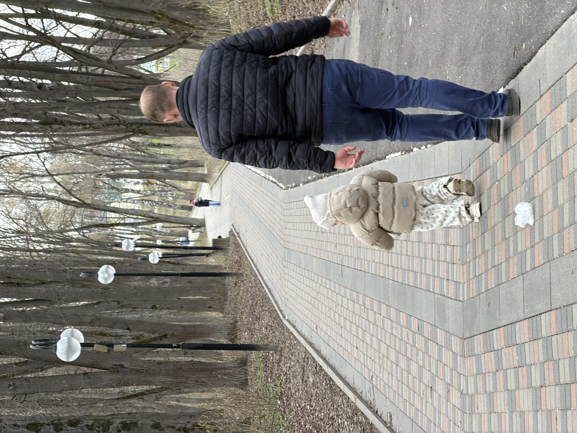 A photograph shows a man and child walking along a brick path in the city of Klin, Russia. The man, seen from the back, wears a black jacket and blue jeans. A child, also seen from the back, wears a beige coat. The path is made of reddish-brown and gray bricks, and it is bordered by trees and streetlights. In the distance, another person is walking.
