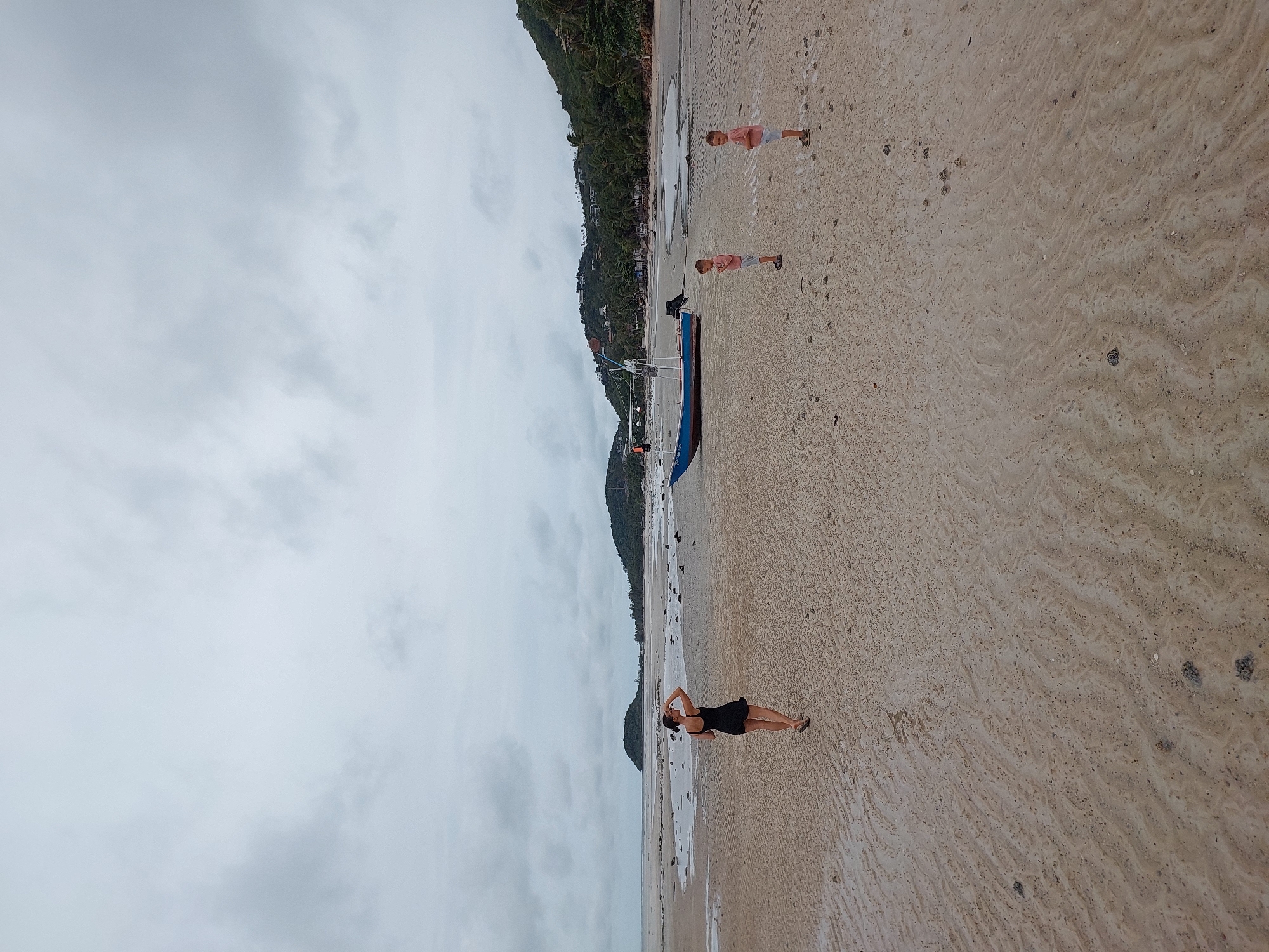 The image depicts a wide, sandy beach on a cloudy day in the Ko Samui District, Thailand. In the foreground, a woman in a black dress stands facing away from the camera, with her hand raised to her forehead as if shielding her eyes from the sun, although the sky is overcast. Further back on the beach, two young boys in pink t-shirts and shorts stand facing each other. A blue and white boat is positioned on the sand between the woman and the boys. The background reveals a tranquil coastline with lush green hills dotted with buildings. The wet sand, marked with ripples and small holes, suggests the tide has recently receded.
