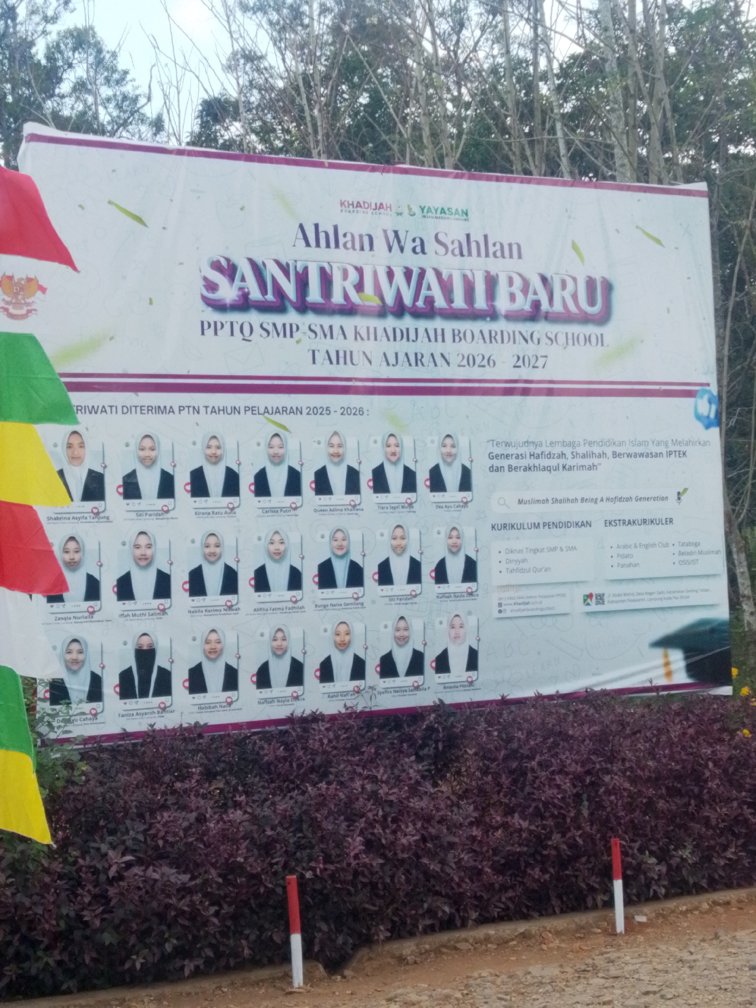 The image is a photograph of a large banner displayed outdoors, likely in front of a school or educational institution. The banner features text and portraits of students. The text on the banner is in Indonesian and appears to be an announcement for a new academic year, listing student names and achievements. The overall theme of the banner is related to Islamic education and boarding school life. The banner is partially obscured by foliage in the foreground. The background consists of trees and a cloudy sky. A small portion of a red and white striped pole is visible in the lower right corner. The specific location of Kurungannyawa, Indonesia is not directly indicated within the image content.