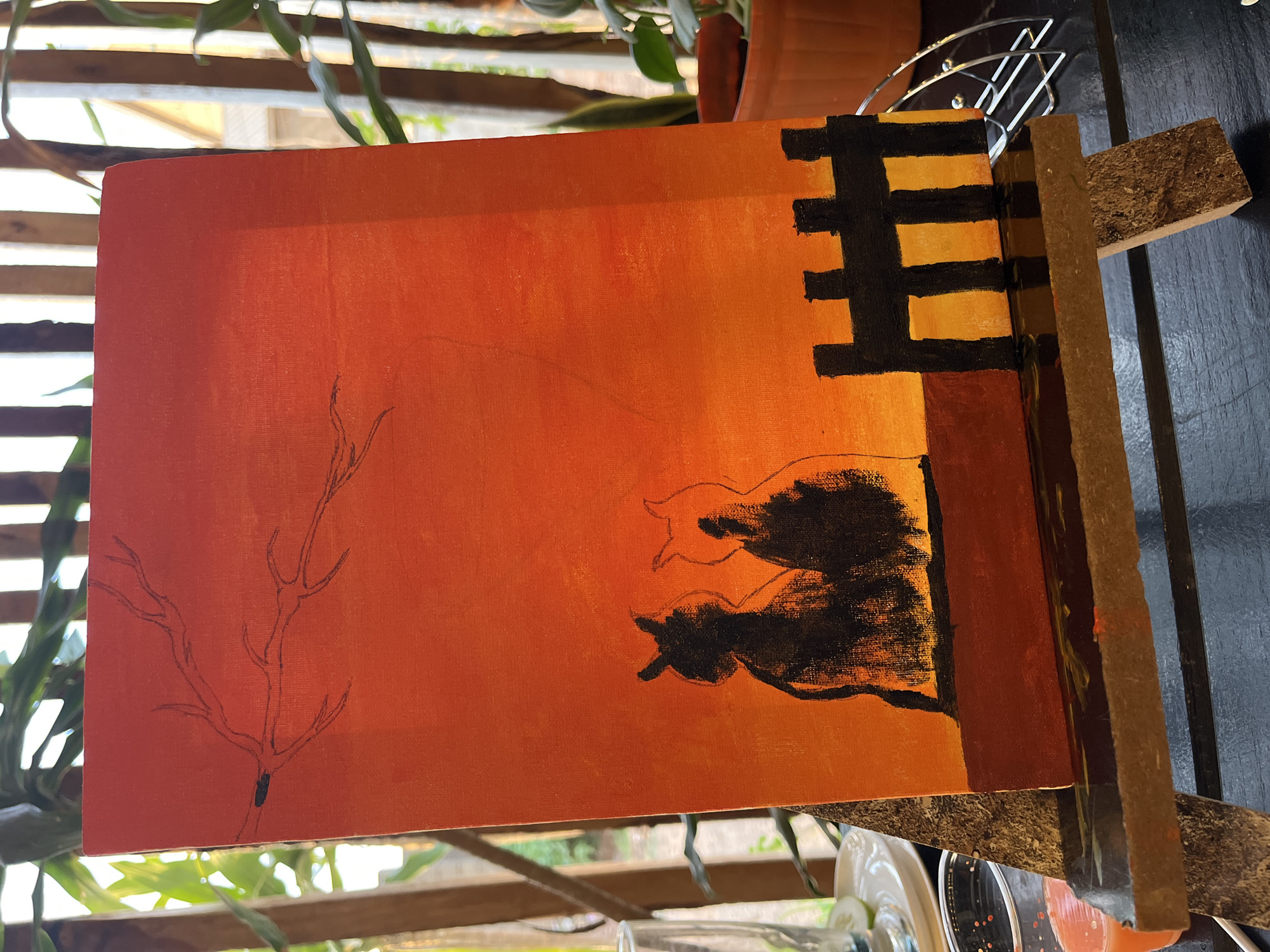 The image shows an unfinished painting on a rectangular canvas, resting on a support. The painting depicts a stylized scene, predominantly in shades of orange and black. On the left, a bare tree branch is drawn in thin black lines. To the right of the branch, two feline figures are rendered in black paint, suggesting the silhouette of cats. Above the cats, a black, grid-like structure resembling a fence or gate stands against a lighter orange background. The canvas itself has an orange base layer. The surrounding environment includes blurred vegetation, wooden slats, and a dark table or counter surface. The exact location within Jos, Nigeria is not discernible from the image.