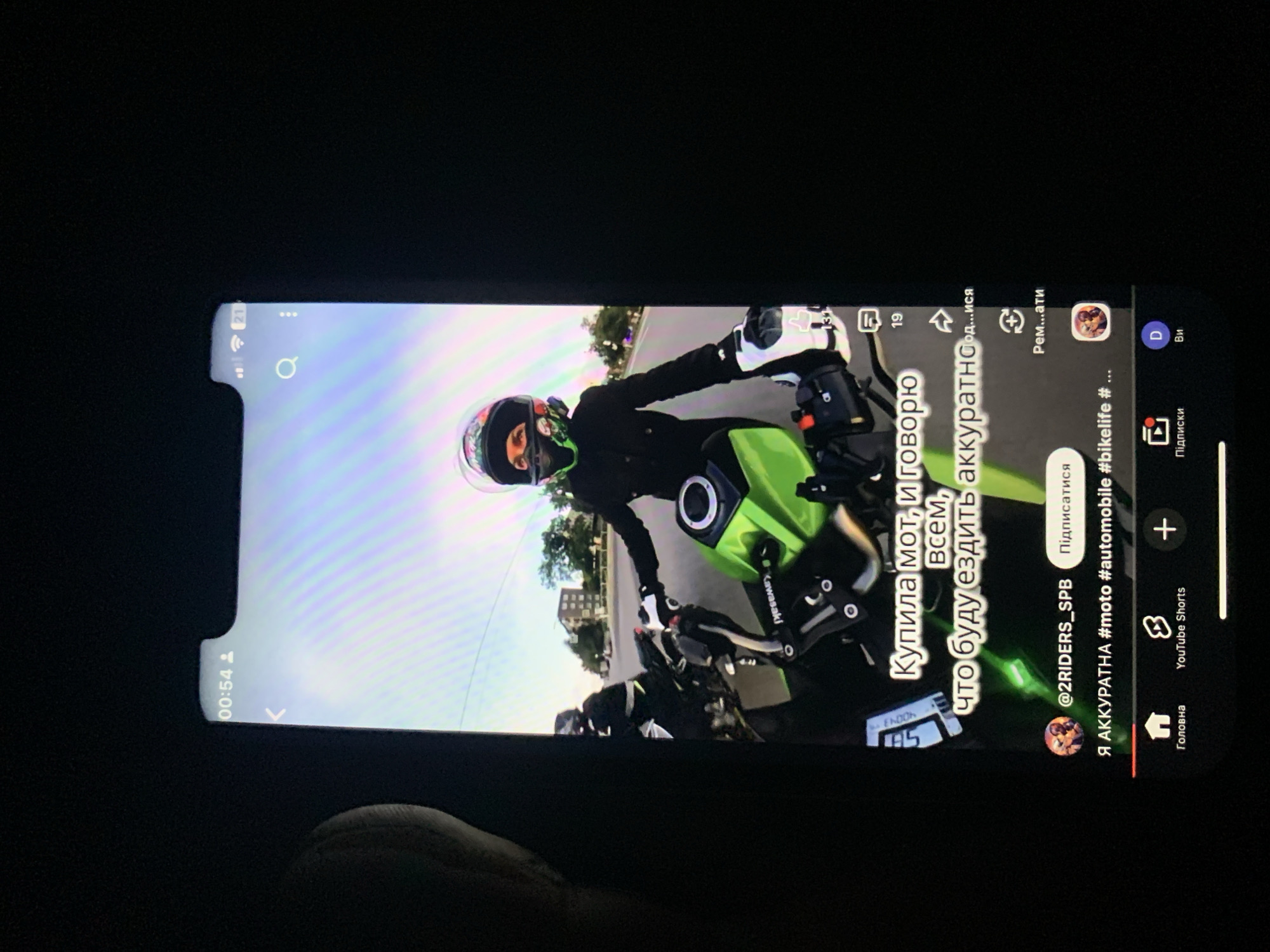 The image displays a vertical screen, likely a smartphone, with a video playing. The video shows a person wearing a motorcycle helmet and riding a green motorcycle. The rider is wearing a black jacket and white gloves. Another motorcycle and rider are partially visible behind the main subject. The background suggests an outdoor environment with buildings and trees, indicative of a city street. Text overlays on the video are in Cyrillic script. The phone's interface elements, such as the time "00:54", battery icon, and navigation bar at the bottom with icons for "Home", "YouTube Shorts", and other options, are visible. The scene appears to be a personal recording or social media content. There is no information in the image to suggest the location is Zhmerynka, Ukraine.