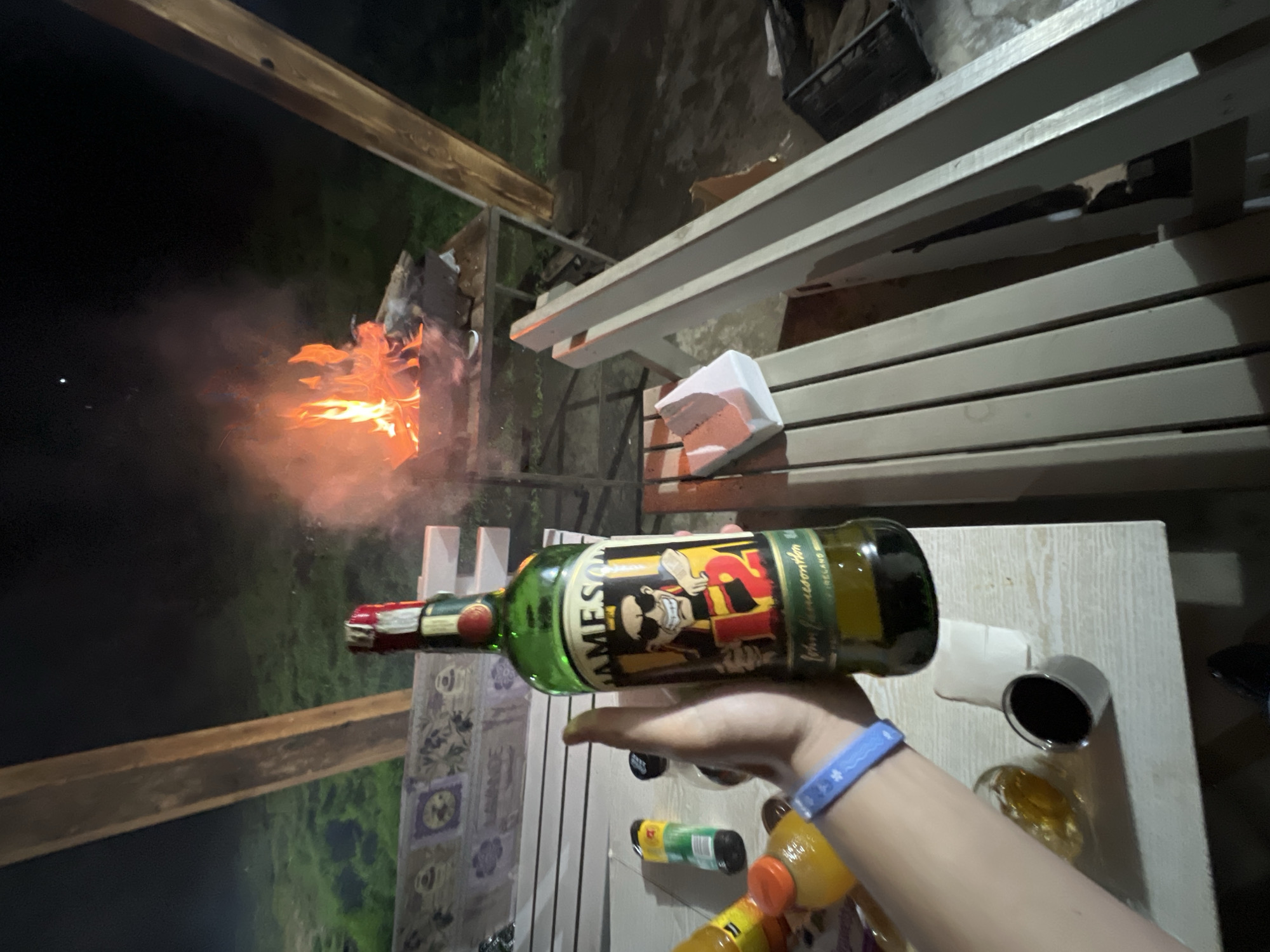 The image depicts an outdoor night scene in Staro Zhelezare, Bulgaria, under a dark sky with a single visible celestial point. In the foreground, a left human hand, wearing a light blue silicone bracelet with white markings, holds a green glass bottle of Jameson Irish Whiskey. The bottle's label features a cartoon character in sunglasses and a hat, with the number "12" prominently displayed. The hand and bottle are positioned above a light-colored wooden slatted table or surface.
On this table, to the left of the bottle, are a clear glass containing a yellowish liquid, a small dark metallic shot glass or measuring cup, a partial view of a yellow plastic bottle with an orange cap, and a green plastic bottle. To the right on the table, a white rectangular object, possibly a tissue pack, rests on a white slatted bench or table extension.
In the mid-ground, a vibrant campfire burns within a metal fire pit or barbecue grill on a stand, casting orange light and producing smoke. The ground surrounding the fire area is dark, appearing grassy and slightly reflective. Architectural elements include two sturdy wooden beams, one angled in the upper left, and another vertical on the right. Behind the table, a decorative wall or structure with patterned tiles, some featuring purple motifs and text ("LAVANDE"), is visible. The scene suggests a casual outdoor gathering or activity.