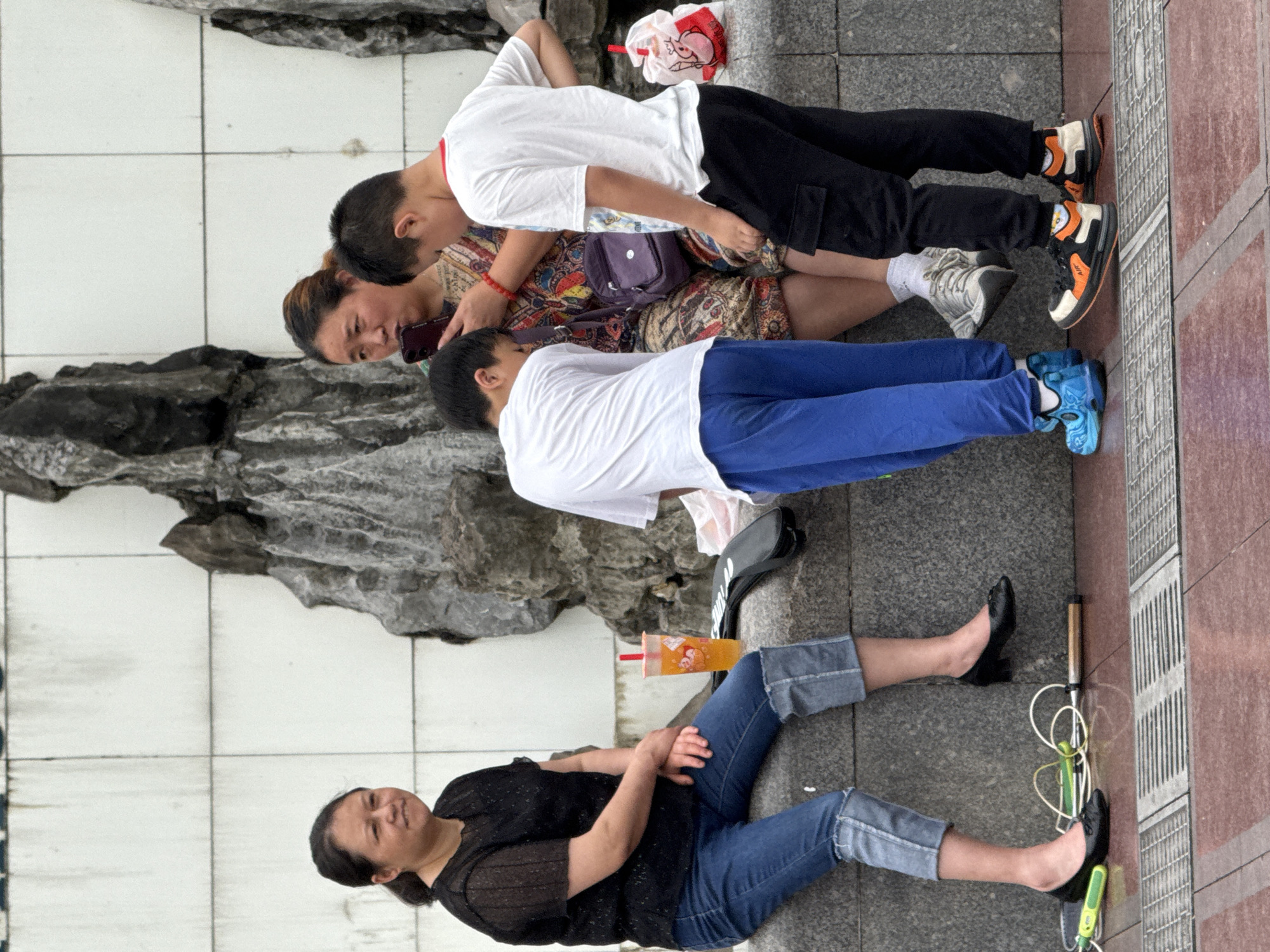 The image shows a group of four people resting on a sidewalk next to a low stone barrier. The location appears to be outdoors, likely within the city of Yuzhong District, China, based on visual cues such as clothing styles, objects, and possible architectural elements.
An adult woman is sitting with her legs outstretched, wearing jeans and a black top. A cup with a straw sits near her. Next to her is another woman, with two boys laying near her, all seemingly related. One boy is wearing a white shirt and dark pants, the other a white shirt and blue pants. The woman near the boys is looking at a phone. Various objects like a bag, sandals, and a jump rope are scattered nearby. The background consists of tiled walls and a possible drainage area, along with what appears to be a stone structure behind the group.