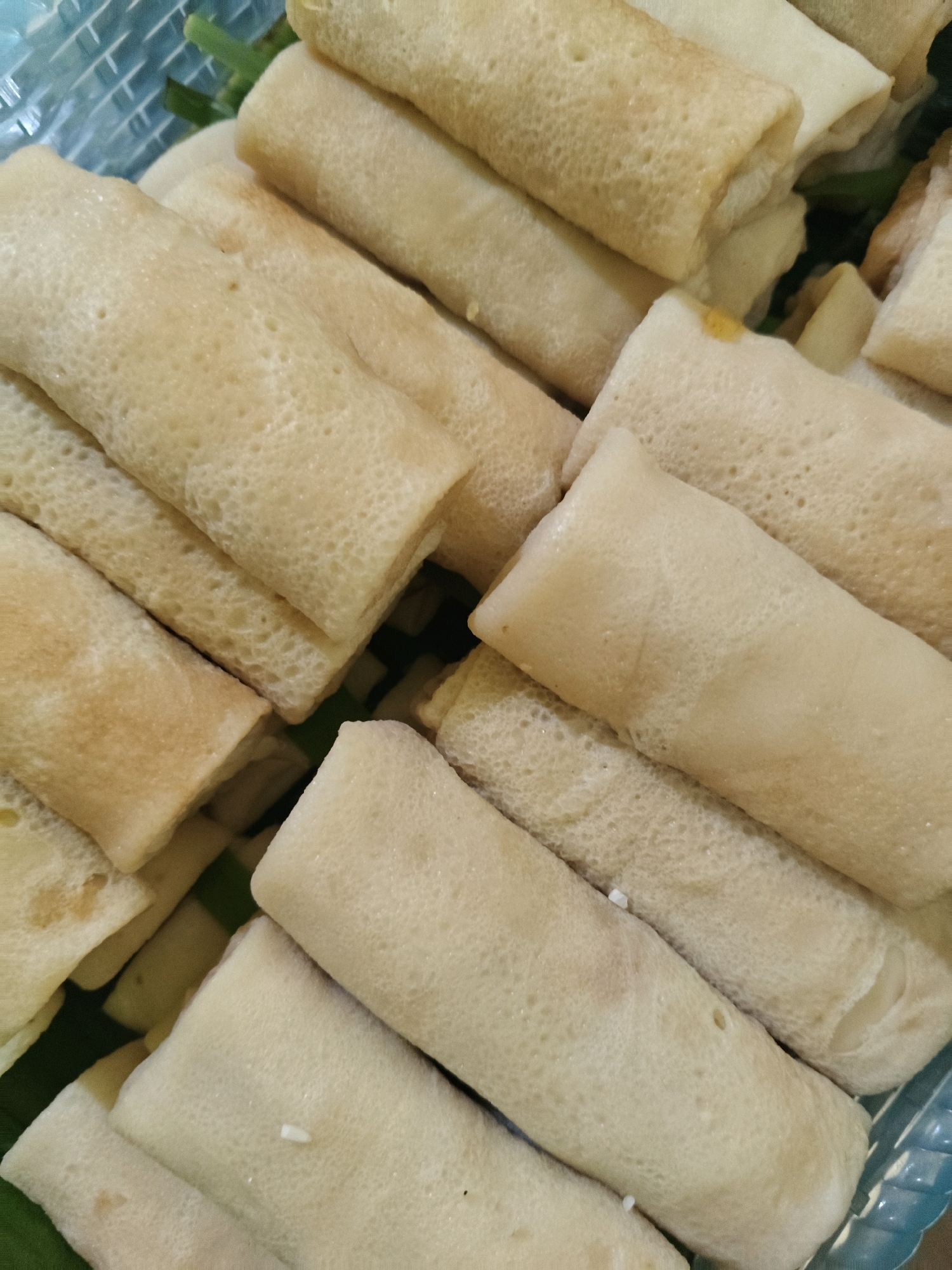 A close-up, overhead shot showcases a blue woven plastic basket filled with a stack of rolled crepes, likely filled and ready for consumption, in Sumberwono, Indonesia. The crepes are a light golden brown and appear to be slightly translucent with visible pores, suggesting they are freshly made. Some crepes have a slight sheen, indicating a soft, pliable texture. The basket's texture is visible in the upper left corner, adding a tactile element to the image. The lighting is soft and diffused, highlighting the food without harsh shadows. The scene is simple and focused entirely on the food item, with no visible people or immediate surroundings beyond the basket.