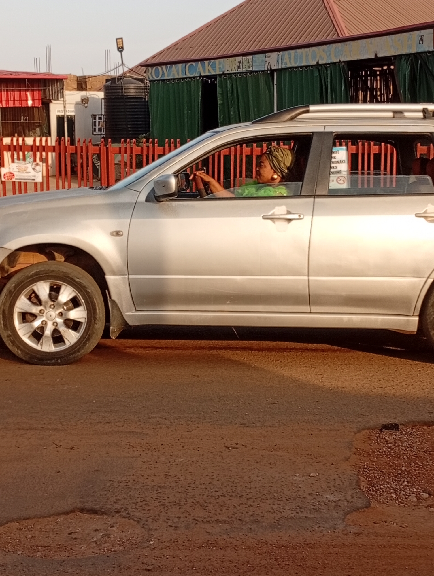 A silver SUV is driving on a paved road in Narayi, Nigeria. The driver is a woman wearing a green headwrap and a green top. A sign on the car window reads "NOT DRINKING ALCOHOLIC DRINKS PENDING." In the background is a building with green curtains and a sign that reads "ROYAL CAKE AUTOS." A red fence and other buildings are visible further in the background. The road is worn and has patches of gravel.