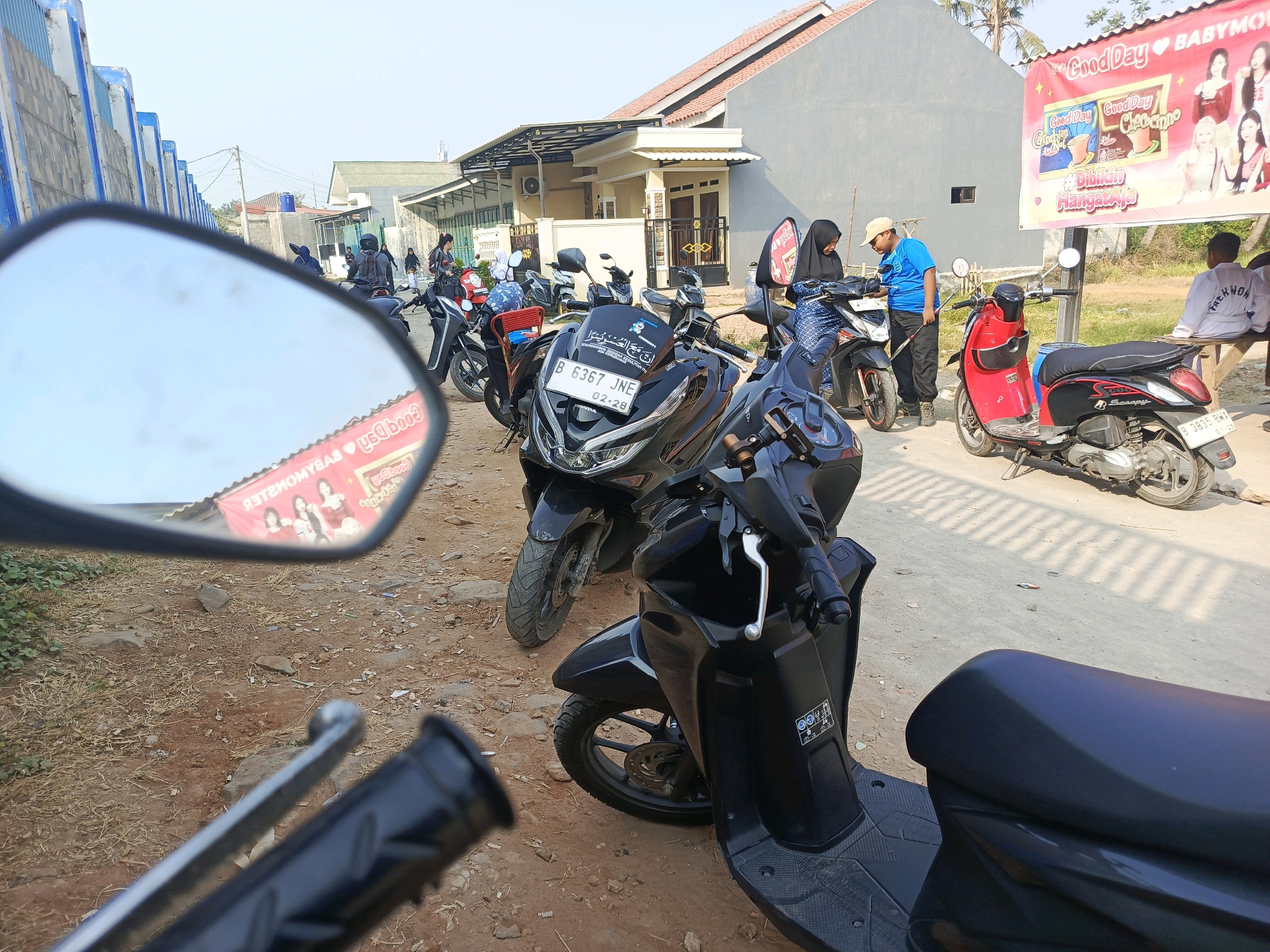 The image depicts a street scene in Jati Mulya, Indonesia, under bright sunlight. Multiple motorcycles are parked on a dirt road. In the foreground, a black scooter is prominently displayed. Behind it, another black motorcycle with a license plate reading "B 6367 JNE" is visible. A red and black scooter with a license plate "B 3835 BWX" is parked further down the street.
Several people are present in the scene. Two individuals wearing hijabs are interacting near a motorcycle. A person in a blue shirt and a cap stands next to them. In the background, a person in a white jacket with "TAEKWON" written on it is seated on a bench. A large banner advertising "Good Day BABYMONSTER" hangs on a building in the background, featuring images of young women and products. The environment includes residential buildings, a blue and white striped wall, and sparse vegetation. The overall impression is a typical street view in a residential area of an Indonesian city.