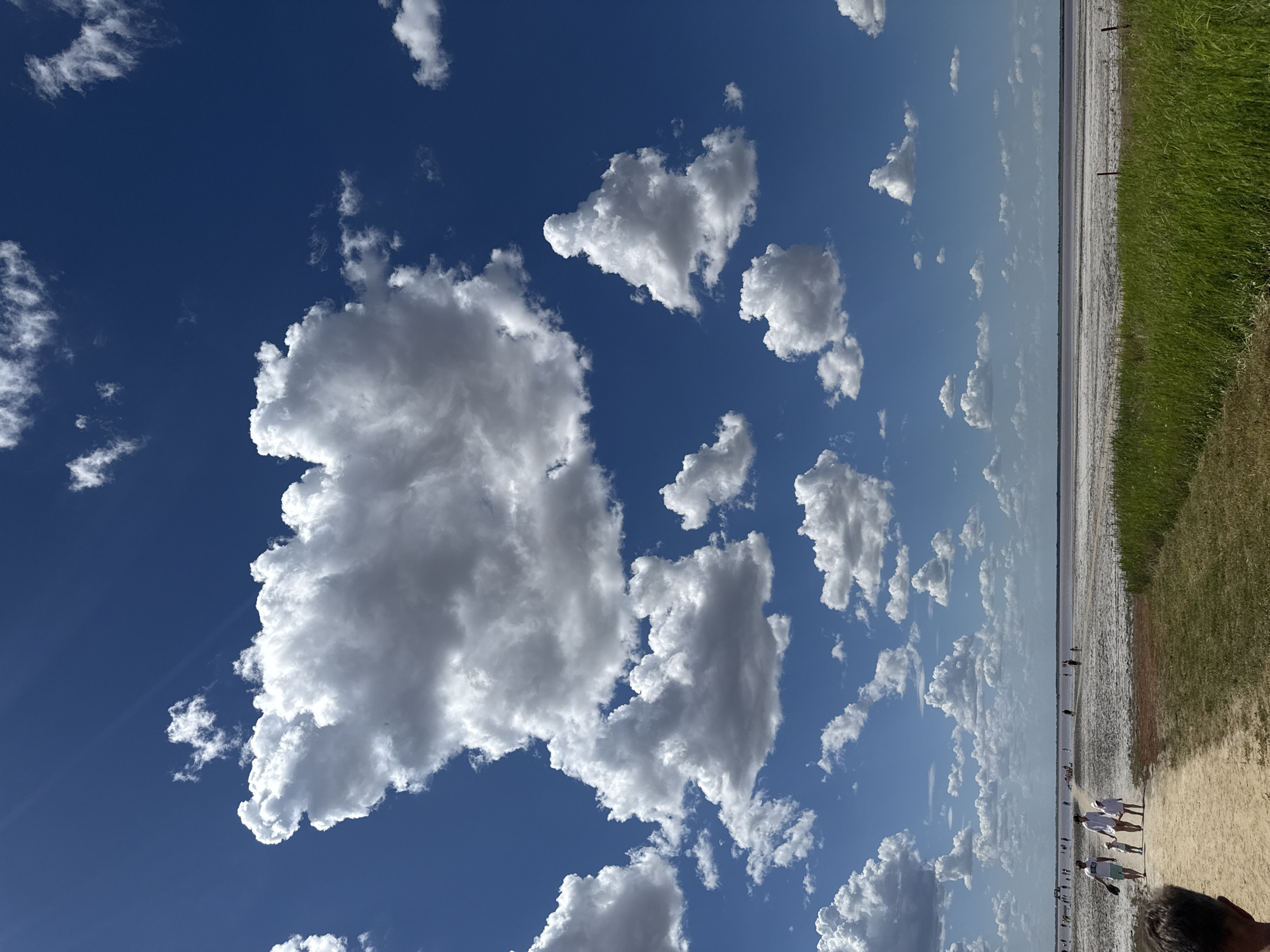 This is a landscape photograph taken on a sunny day at a salt lake in Bursol, Russia. The sky is a deep blue with numerous white, fluffy cumulus clouds scattered across it. The horizon is marked by a vast, flat expanse of what appears to be a salt lake, with a dry, textured surface interspersed with pinkish hues in the distance. Along the shore, a few people are visible, walking and standing. In the foreground, a dirt path leads towards the lake. To the right, a patch of vibrant green grass grows. The scene is peaceful and evokes a sense of open space and natural beauty.