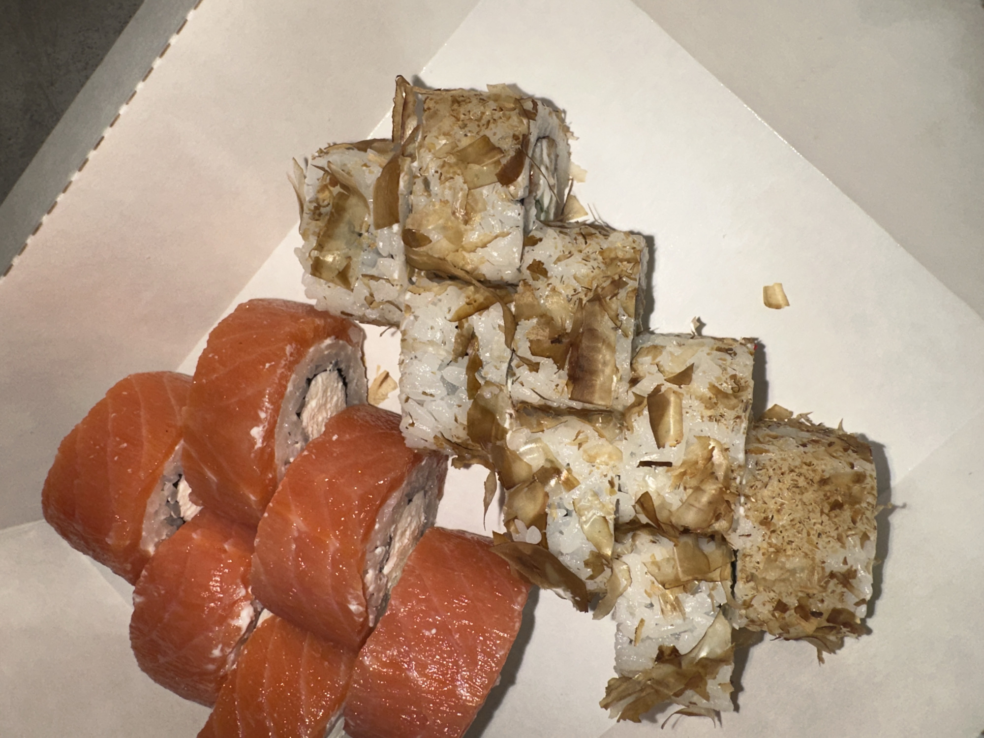 A top-down shot depicts an assortment of sushi rolls in a white takeout container. On the left, a row of sushi rolls is visible, wrapped in salmon. To the right, there is another set of rolls covered in what appears to be dried fish flakes. The takeout container has a slightly visible angular edge. Based on geotags, this photo originates from the city of Chaikovsky, Russia.