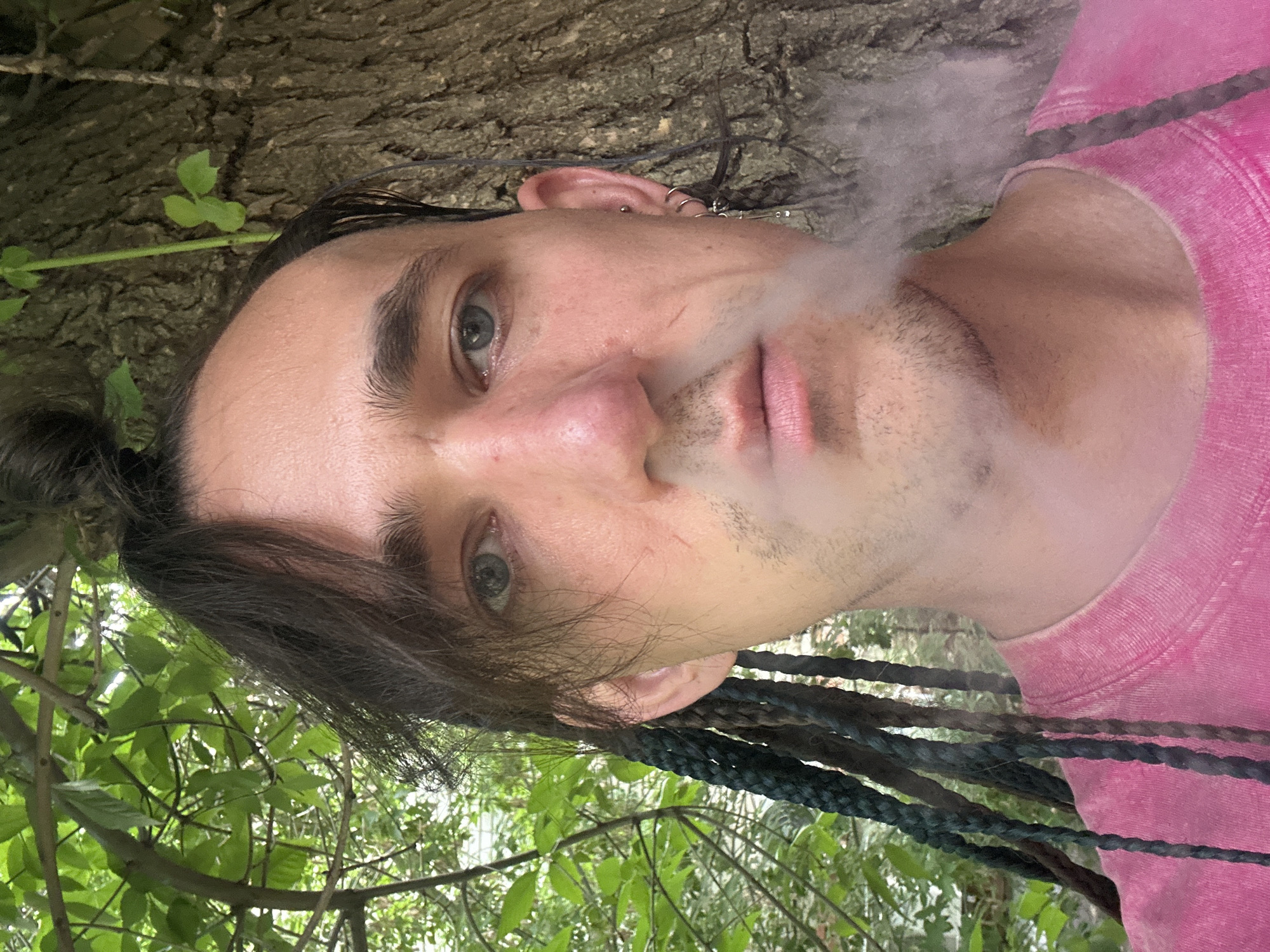 This is a close-up, self-portrait photograph of a young person in an outdoor setting. The individual has light skin, blue-green eyes, and dark hair styled in braids that hang down. They are wearing a bright pink t-shirt and appear to be exhaling a cloud of smoke, suggesting they are smoking or vaping.
The background is predominantly green foliage, indicating a natural environment, possibly a park or wooded area. A textured tree trunk is also visible behind the subject's head, further reinforcing the outdoor, natural setting. The lighting suggests it is daytime, with the sunlight dappling through the leaves. There are no visible signs of text within the image. The overall emotion conveyed by the subject's expression is neutral.