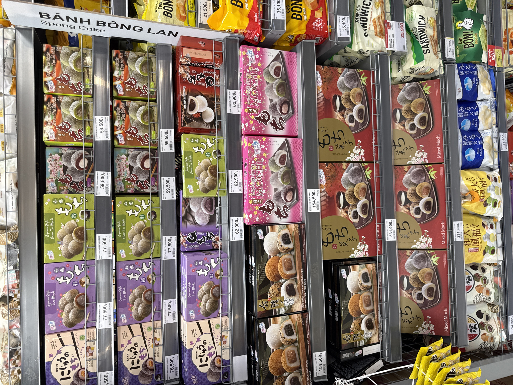 The image shows a well-lit retail store aisle, likely a supermarket, in Nha Trang, Vietnam. The shelves are stocked with a variety of Japanese mochi products in colorful packaging. The products are primarily displayed in boxes and bags, with varying flavors and styles such as "Taro Mochi," "Green Tea Mochi," and "Mixed Mochi." Some packaging features imagery of the mochi itself, while others use decorative elements like cherry blossoms and Japanese calligraphy. Prices are clearly visible on small tags below many of the products. The overall environment is clean and organized, typical of a modern retail setting. There are no people visible in the image.