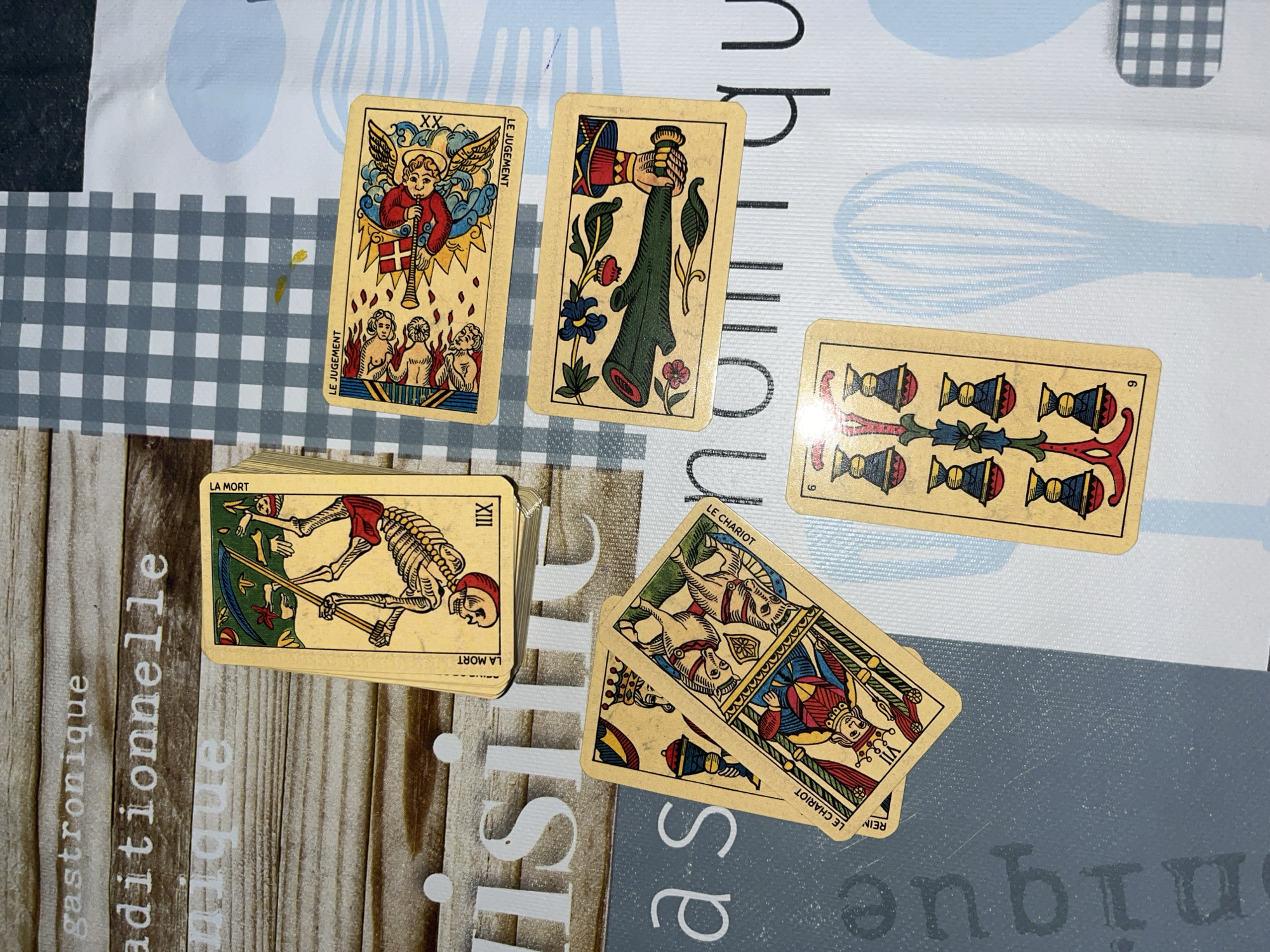 The image shows a tarot card reading spread out on a tablecloth. There are five tarot cards visible: "Le Jugement," "6 de Coupe," "La Mort," "Le Chariot," and one other card. The environment includes elements of "gastronomique aditionelle nique" on the tablecloth. The scene is not explicitly linked to Santeru/San Vero Milis, Italy, or any city. There are no people visible in the image.
