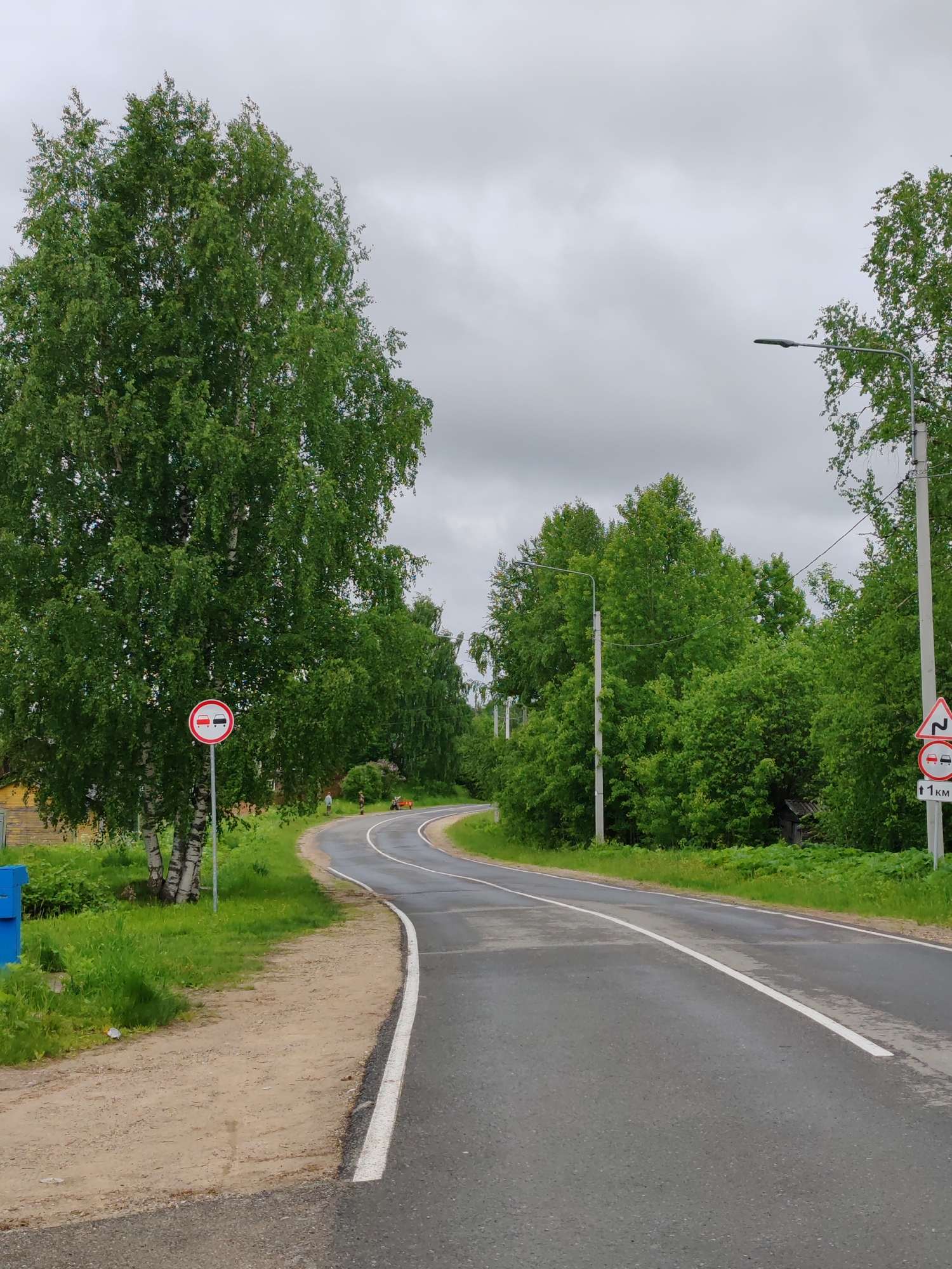 A rural road in Чухлэм, Russia, curves gently to the right under an overcast, cloudy sky. The asphalt surface of the road appears wet, suggesting recent rain. A continuous white line marks the right edge of the road, while a dashed white line indicates the center or left edge, leading around the bend.
On the left side of the road, a large birch tree with distinctive white bark and dense green foliage stands prominently. Below it, a circular regulatory sign with a red border displays two cars (one red, one black/grey) facing each other, indicating "No Overtaking." Further left, a blue box-like object is visible near the ground. A dirt shoulder runs alongside this part of the road.
In the distance, on the left side of the bend, two small figures are visible. One appears to be standing, while the other seems to be near a small red wheeled object, possibly a cart or small piece of equipment. Behind the birch tree, a glimpse of a weathered wooden building can be seen.
On the right side of the road, lush green trees and bushes line the path, forming a dense wall of foliage. A utility pole with a streetlamp extends from the right, and another lamp post is visible further down the road. Attached to the utility pole is a white triangular warning sign with a red border, depicting a winding road symbol (two sharp turns). Below this, another sign shows two cars (red and black) facing each other, and below that, the text "↑1 KM," indicating "No Overtaking for 1 Kilometer."
The scene portrays a quiet, green landscape on a damp, cloudy day.