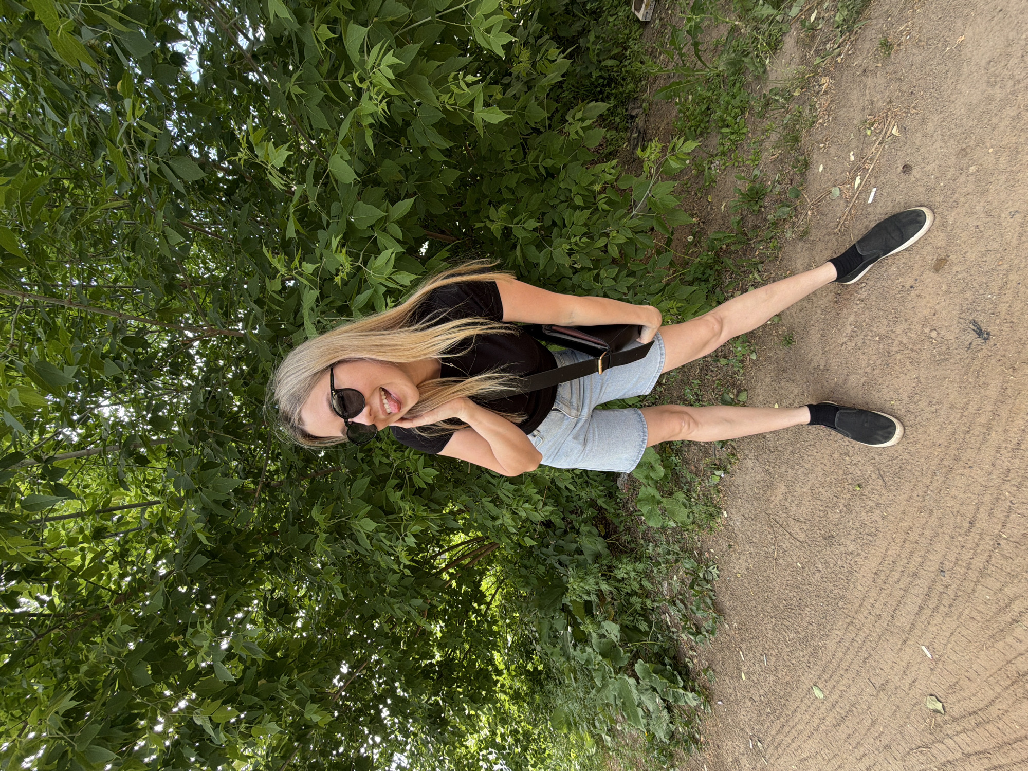 A young woman with long blonde hair is standing on a path in Chaikovsky, Russia. She is wearing sunglasses, a black t-shirt, and denim shorts. She is holding a black purse with a strap. She is sticking out her tongue and smiling. The path is dirt and has some grass and leaves on it. There are trees and bushes in the background.