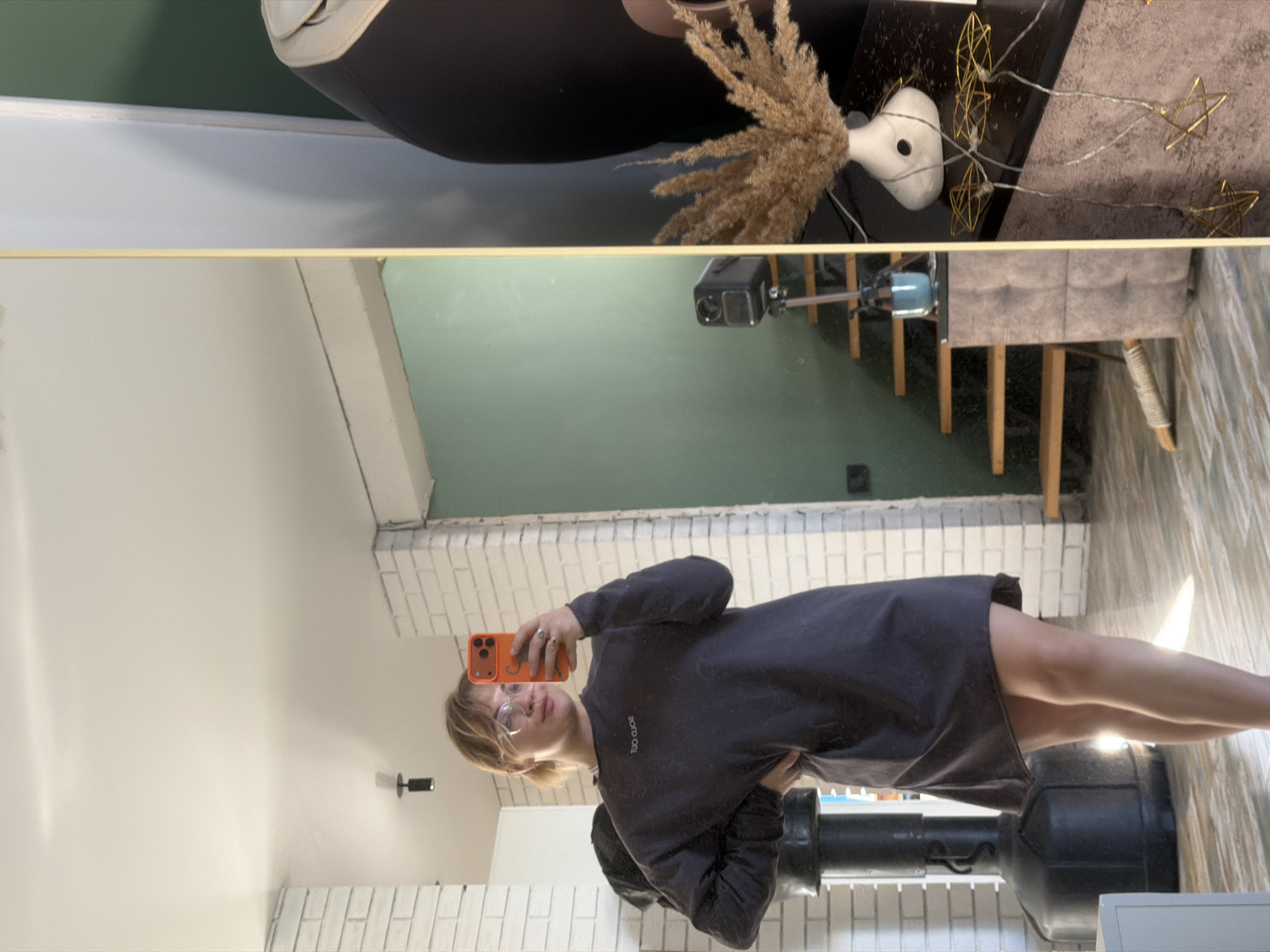 Here is a concise description of the image:
The image appears to be a selfie taken in a mirror. A woman with short blonde hair and glasses is holding an orange phone. She is wearing a dark gray or black long-sleeved shirt or dress. The mirror reflects a room with white brick walls and a green wall. In the background, there's a heavy bag.
The upper part of the mirror shows a surface with decorative elements, including a vase of wheat, string lights, and a gray surface, likely from another room or a reflection of items in the mirror's immediate vicinity.
The location is Большая Шильна, Russia, based on available information.