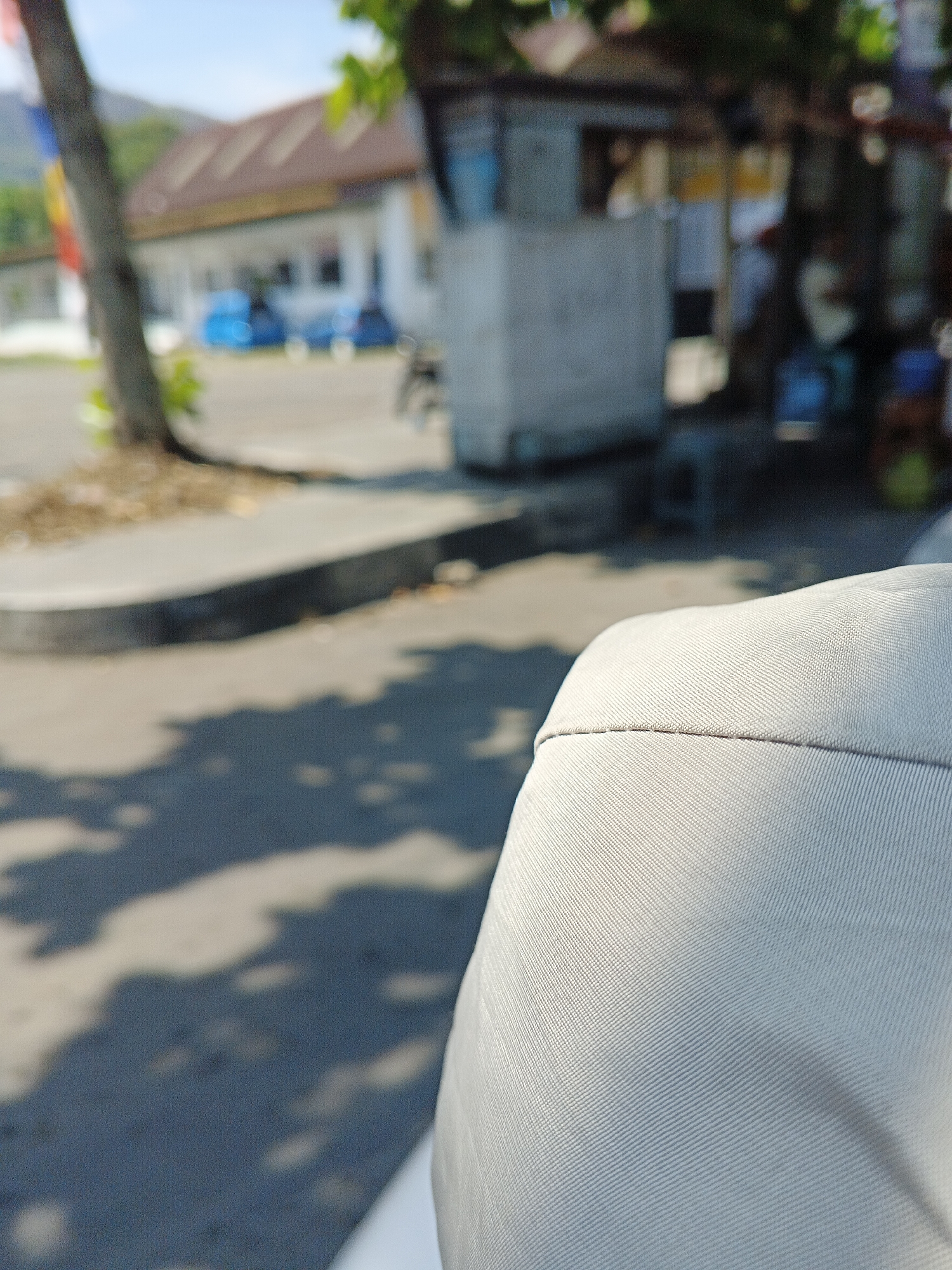 The image shows a close-up of a light-colored fabric, likely upholstery or clothing, in the foreground. The background is blurred, suggesting it is out of focus or that the photograph was taken from a moving vehicle. The background scenery includes a tree trunk on the left, a building with a brown roof and white walls, some blue objects that might be vehicles, and a concrete structure. The scene appears to be outdoors, possibly in a public area such as a street or parking lot, with dappled sunlight casting shadows on the ground. The location is identified as Sukabumi, Indonesia, suggesting an urban or semi-urban environment. There are no distinct individuals or specific actions readily identifiable due to the blurriness of the background.