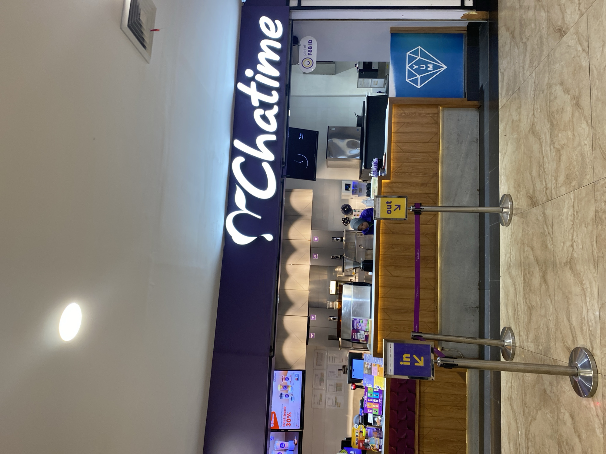 The image shows the exterior of a Chatime store, a popular bubble tea franchise, located in Banda Aceh, Indonesia. The store's prominent purple and white signage, featuring the "Chatime" logo, is clearly visible at the top of the establishment. In front of the counter, stanchions with purple ropes guide customers, with a yellow sign indicating "in" with an arrow pointing to the right and another sign indicating "out" with an arrow pointing to the left.
The counter area is well-lit, with various equipment and supplies visible, including coffee machines, blenders, and product displays. A cashier, partially obscured by the counter, appears to be attending to a customer. Behind the counter, shelves are stocked with drinks and other merchandise.
The flooring in front of the store is a light-colored marble pattern, and the walls have a textured finish. The overall impression is of a clean and modern retail space designed to attract customers. The presence of the "part of F&B ID" logo suggests that this Chatime store is part of a larger food and beverage network in Indonesia.