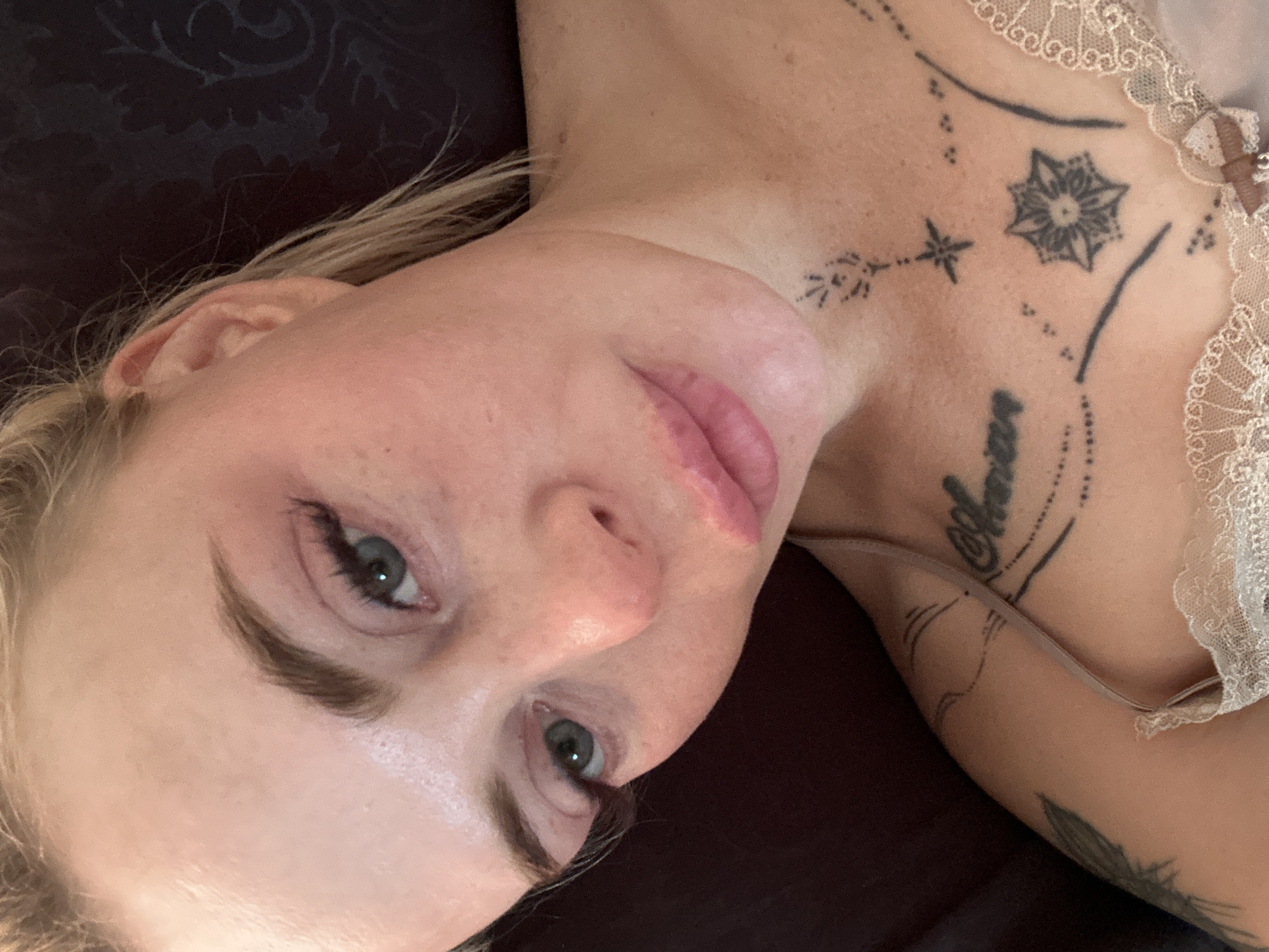 A woman with blonde hair and green eyes is shown from the chest up. She is wearing a light-colored lace camisole. She has several tattoos visible on her chest, neck, and shoulder, including a floral design on her chest and what appears to be lettering on her collarbone. Her makeup includes mascara and lipstick. The background is dark and textured.