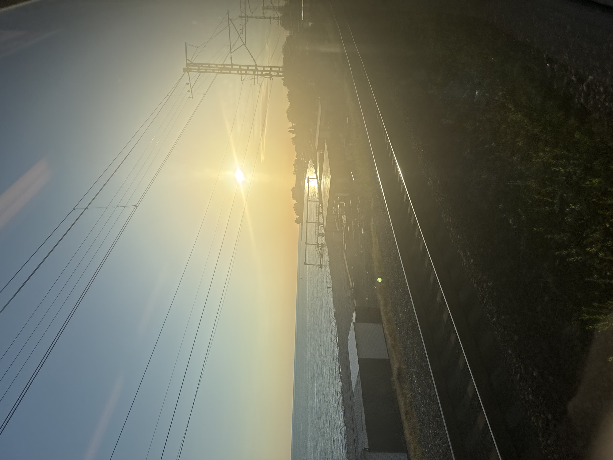 The image depicts a vast, sun-drenched landscape featuring railway tracks, a large body of water, and power lines. The time of day is clearly either sunrise or sunset, indicated by the intense, low-angled sun and the warm, golden light bathing the scene.
The sun is the most prominent element, positioned centrally on the right side of the frame (when correctly oriented), casting a brilliant glare that creates significant lens flare and washes out parts of the sky and water. The sky transitions from a soft blue on the left to a vivid yellow and orange closer to the sun.
Multiple dark power lines crisscross the upper portion of the sky, leading to large, industrial-style pylons visible on the distant horizon. Below the horizon, a wide expanse of water, likely a lake or sea, stretches out, reflecting the sun's intense light. Along the distant shoreline, faint outlines of land, possibly with some trees or structures, can be discerned.
In the foreground, two parallel railway tracks run horizontally across the bottom of the image, flanked by dark, rocky ground and patches of green vegetation. Several indistinct, light-colored rectangular structures are visible close to the tracks. The perspective suggests the photograph was taken from a moving vehicle, possibly a train, looking out a window.
The scene conveys a sense of peaceful travel during the "golden hour," with clear weather and a serene atmosphere. No visible text is present in the image.
