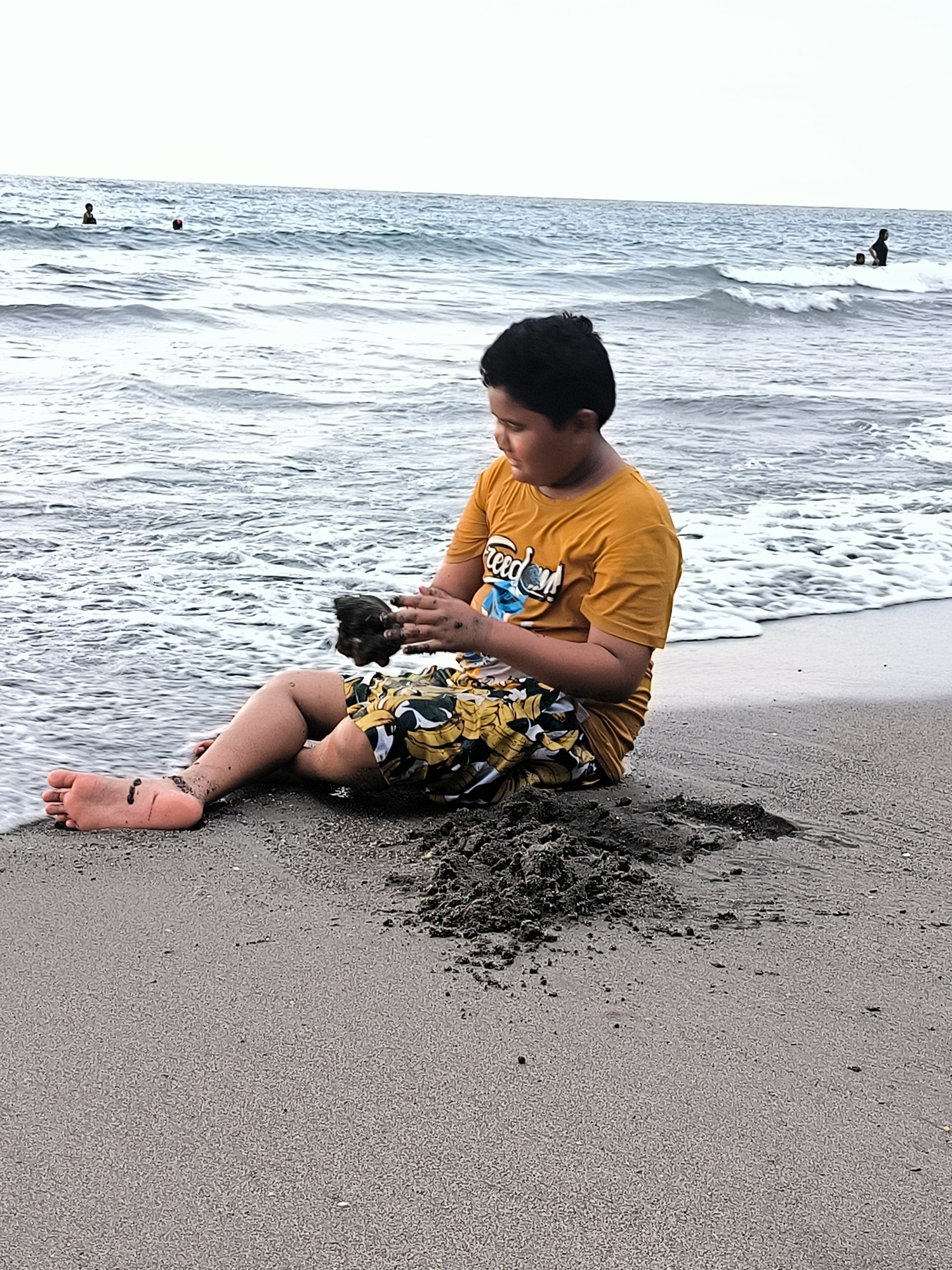 The image presents a coastal scene in Lhokseumawe, Indonesia, depicting a young individual on a sandy beach with the sea in the background. In the foreground, a person, identified as a child or young adolescent, sits on the greyish-brown sand at the water's edge. This individual has dark hair and wears a mustard yellow short-sleeved t-shirt with a partially visible blue graphic and patterned yellow and black swim shorts. The person is actively engaged in handling a dark, wet mass of sand or mud with both hands, looking down at the activity. A pile of similar dark, wet sand is positioned directly in front of the individual on the beach. Bare feet, coated with sand and exhibiting dark smudges, are visible. The background reveals the sea under an overcast sky, characterized by greyish-blue water with gentle waves breaking on the shore. Multiple figures are present in the water at varying distances: one individual is partially submerged on the far left, another person wearing a red head covering is further out in the water, and two individuals, appearing to be an adult and a child, are observed in the breaking waves on the right side of the frame.
