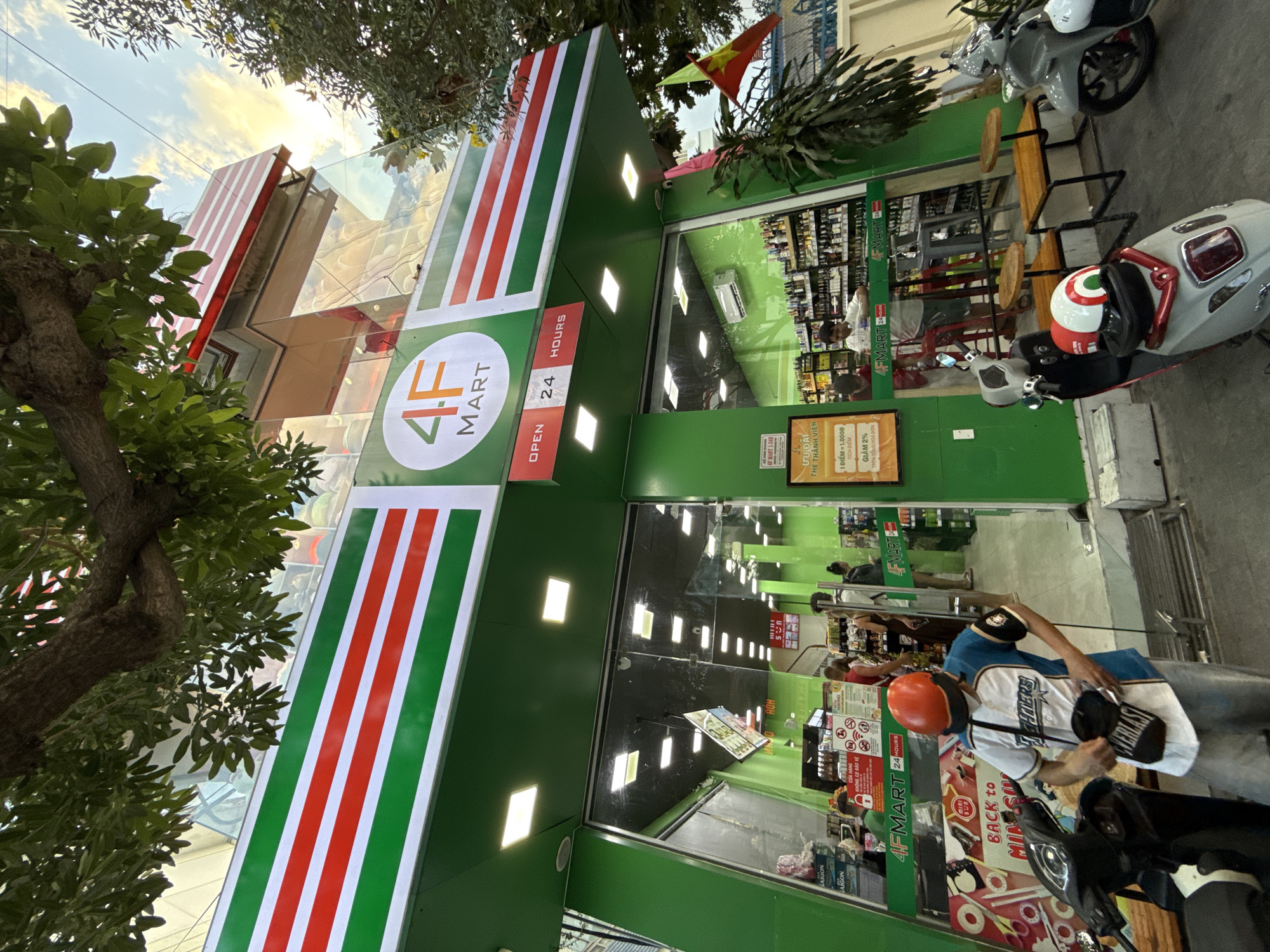 The image depicts a brightly lit, modern convenience store named "4F MART" situated on a street in Nha Trang, Vietnam. The store's facade is dominated by vibrant green with horizontal stripes of red and white. A large sign above the entrance reads "OPEN 24 HOURS". Inside, shelves are stocked with various products, including drinks and snacks, and several customers are visible browsing. Two men are standing at the counter, and a woman is looking at products on the shelves. Outside the store, a person wearing an orange helmet and a baseball jersey is standing near a parked black scooter. Another scooter, light gray in color, is parked to the right. A small Vietnamese flag is displayed prominently on a pole to the right of the store entrance. The sky above is partly cloudy, suggesting it is either late afternoon or early morning. Trees with dense green foliage frame the upper portion of the image.