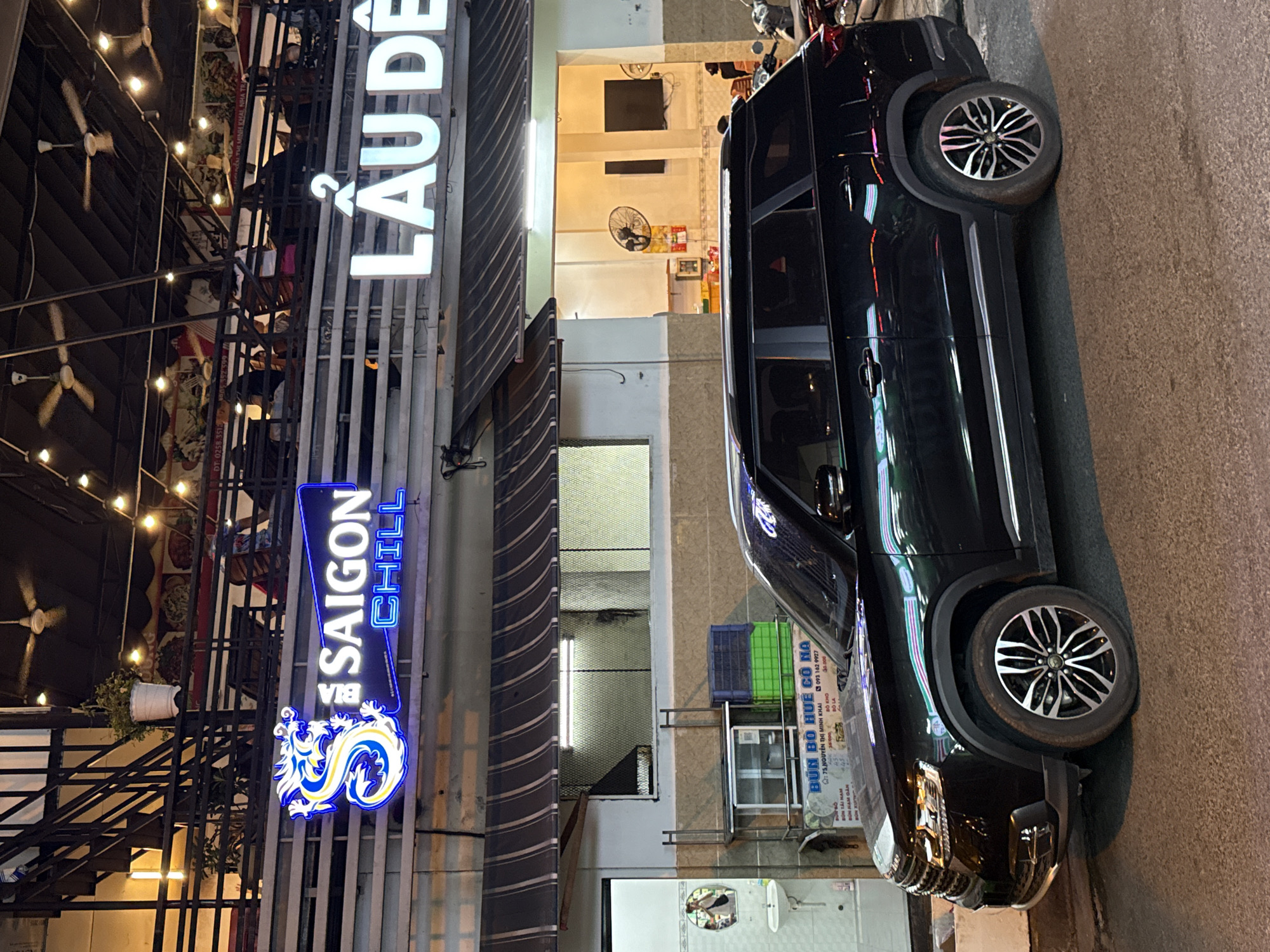 The image shows a black SUV parked on a street at night in front of a building with multiple levels. A neon sign on the building reads "SAIGON CHILL" with a dragon design. Another sign above it reads "LẨU DÊ". The building appears to be a restaurant or food establishment. There are people visible on the upper levels of the building. The street is paved, and there are other vehicles, including motorcycles, parked further down the street. The overall environment suggests an urban setting in Vietnam, likely Nha Trang given the context.